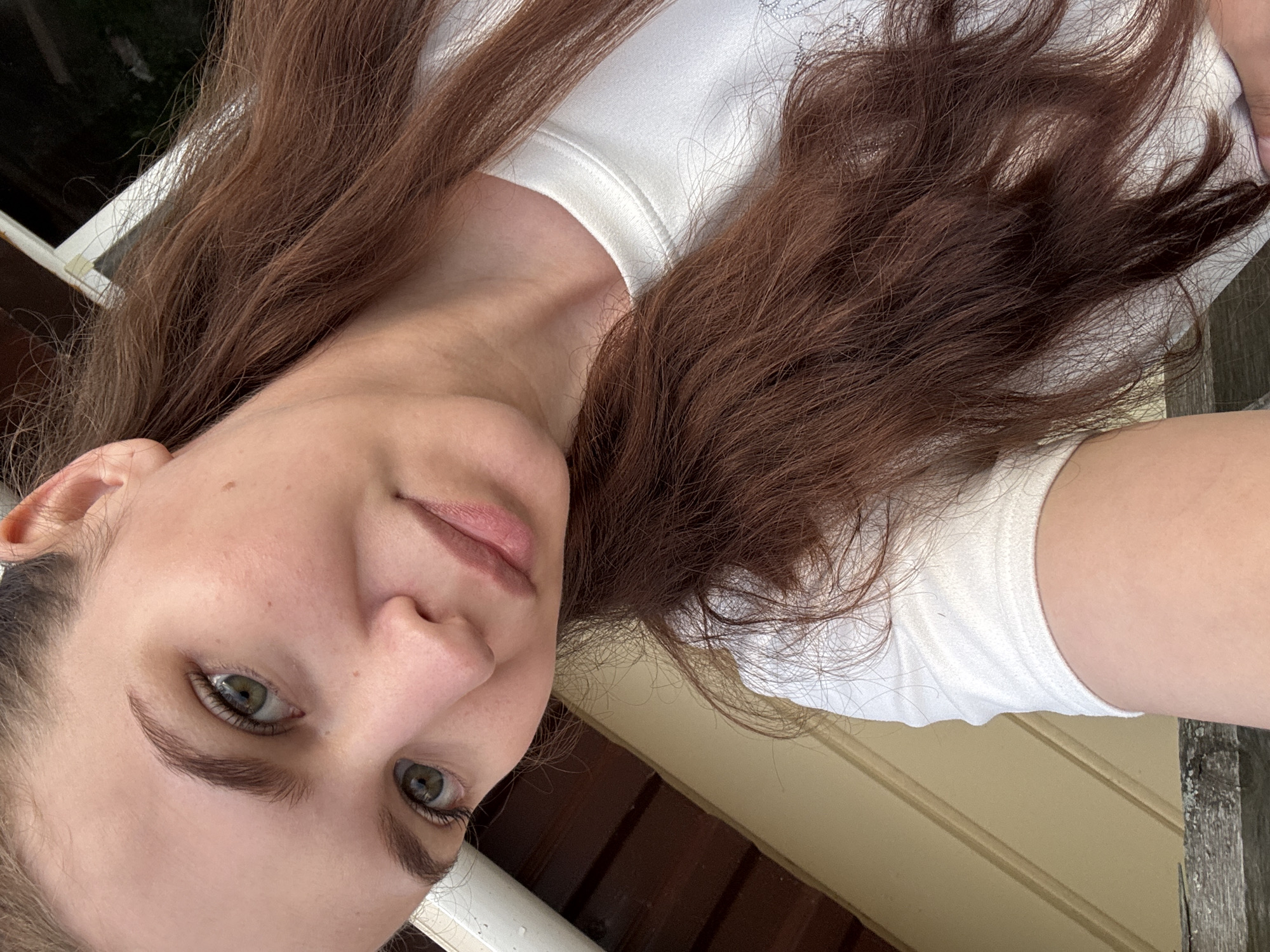 A young woman with auburn hair is taking a selfie outdoors. She is wearing a white t-shirt and her head is tilted, with her face filling most of the frame. Her green eyes are looking towards the camera, and she has natural-looking makeup. Behind her, a wooden structure with horizontal planks is visible, along with a white railing and some greenery. The overall lighting suggests it is daytime. The location is the city of Nizhnyaya Gar', Russia.