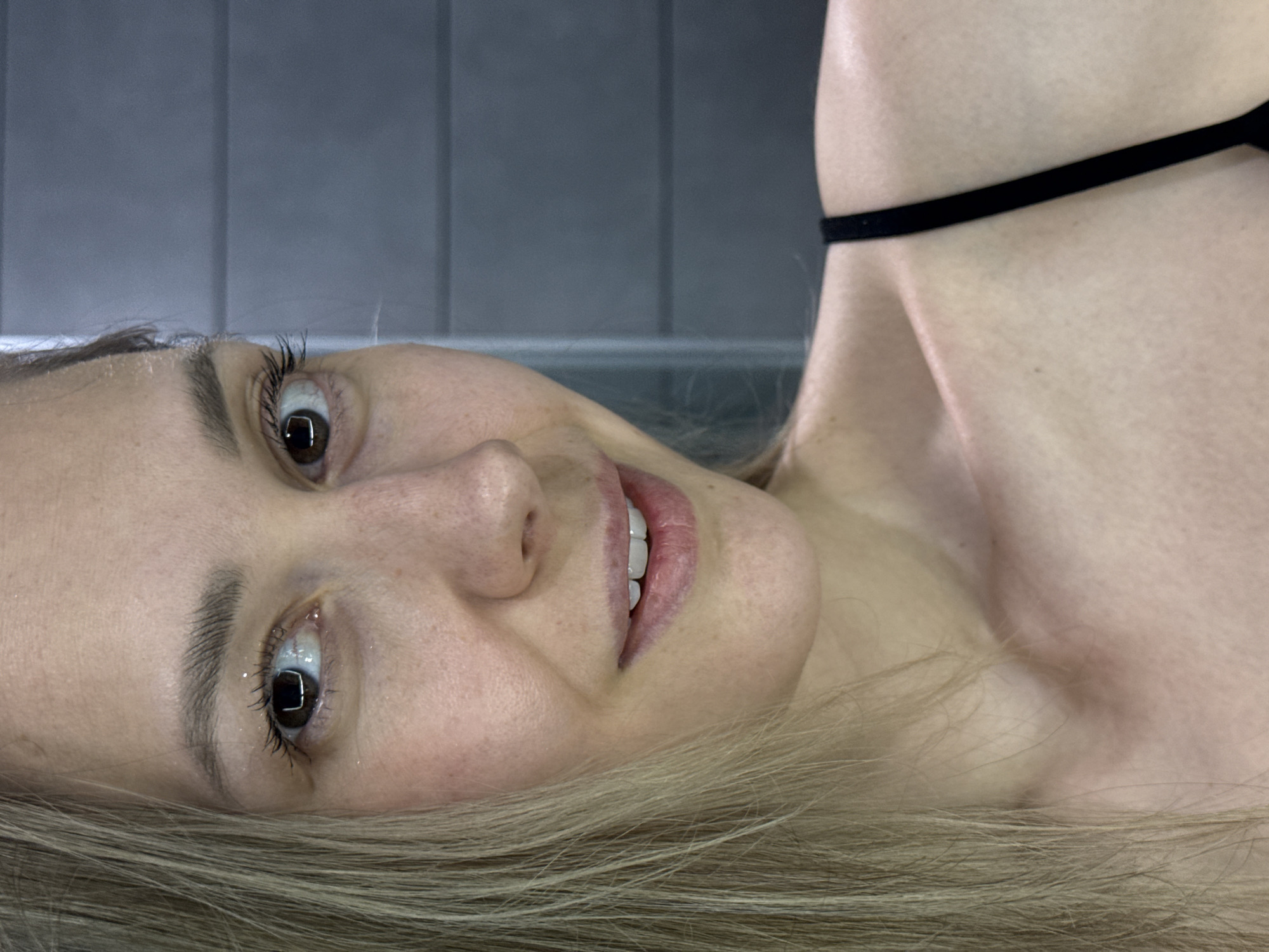 A woman with fair skin and light brown hair is lying on a surface, facing the camera with a slight smile. She has brown eyes and appears to be wearing minimal makeup. A thin, black strap is visible across her upper chest. The background consists of gray, vertically oriented panels. There is no specific information to confirm the location in the city of Chaikovsky, Russia.
