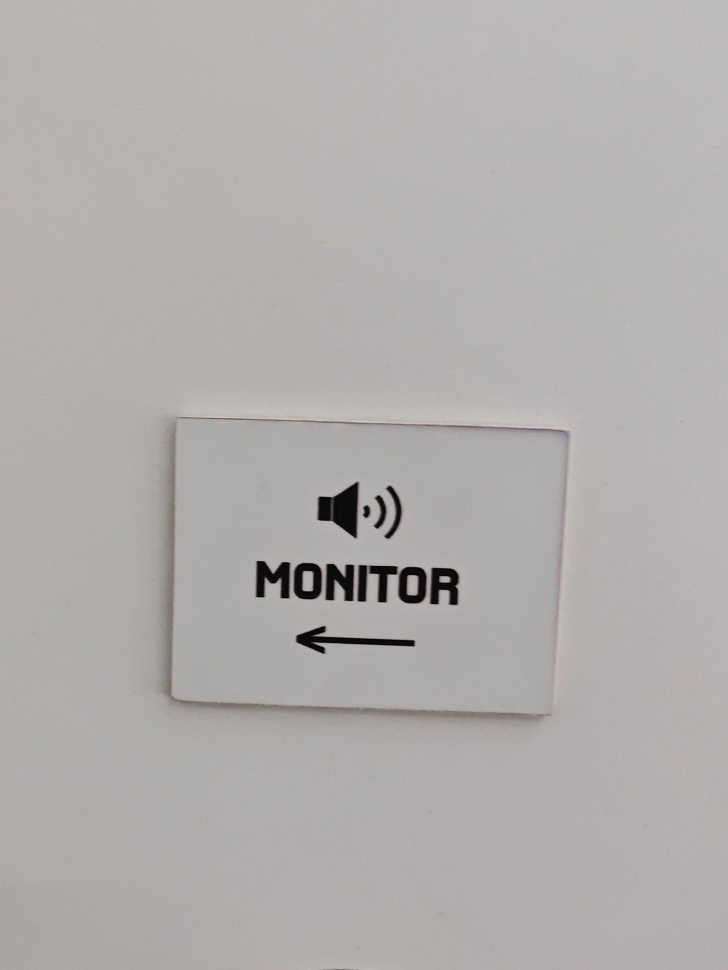 A white rectangular sign with black lettering and a symbol is affixed to a pale gray wall. The sign displays a black icon of a speaker with sound waves emanating from it, positioned above the word "MONITOR" in capital letters. Below the word "MONITOR," a black arrow points to the left. The sign is oriented horizontally. The wall appears to be a neutral interior surface, possibly a wall in a public building or office. No people or other objects are visible in the immediate vicinity. The image does not provide any information that would allow for the determination of its location in the city of Turin, Italy.