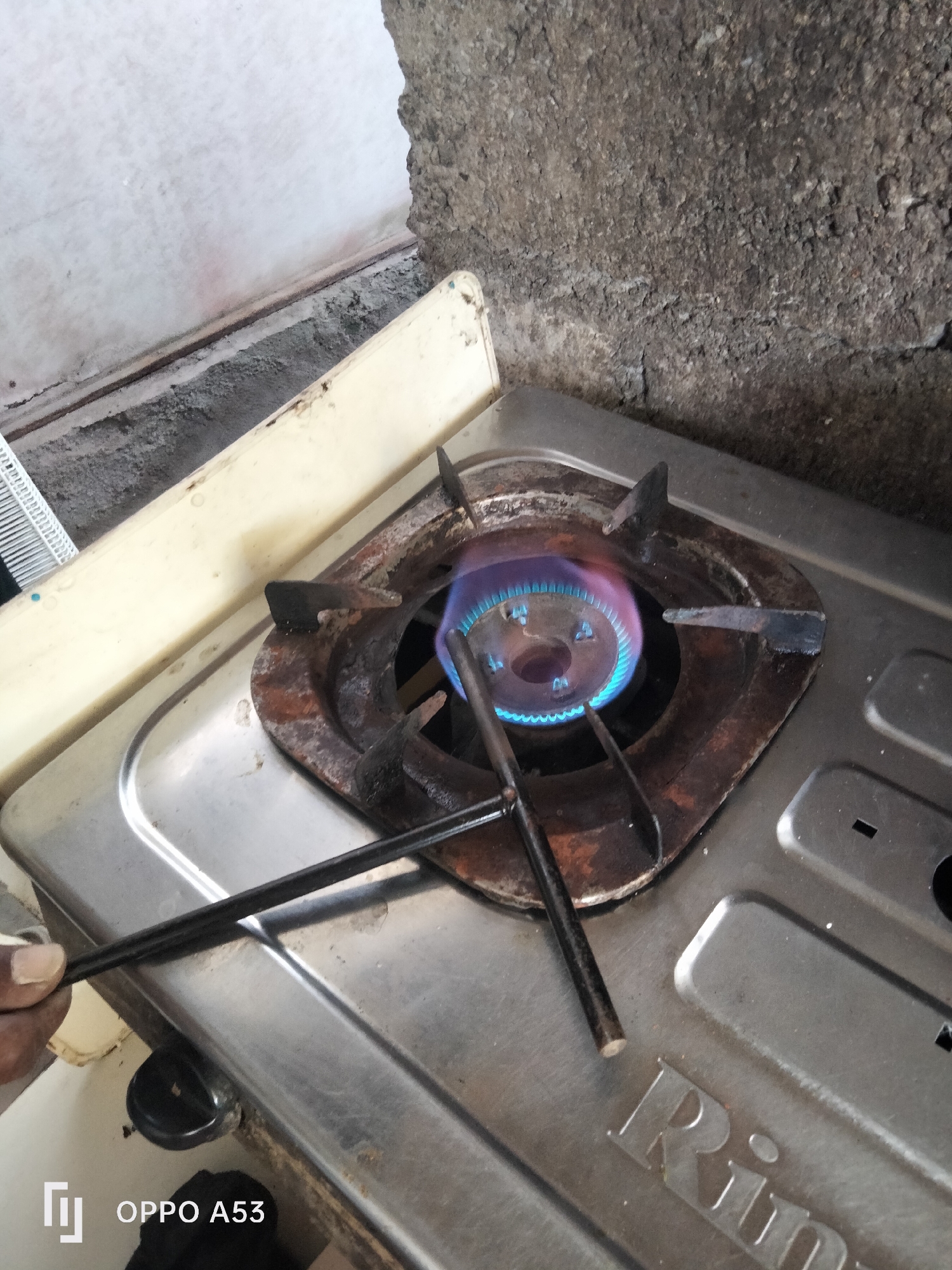 This is a close-up, eye-level, outdoor shot of a gas stove burner with a blue flame igniting. The stove appears to be made of stainless steel and has some rust around the burner. Two black chopsticks are being held over the flame by a person's hand, suggesting an attempt to cook or light something. In the background, a rough, gray concrete wall is visible. The bottom of the image shows a portion of the stove with the brand name "Rin" visible. The presence of the gas stove and the materials suggest a residential or food stall setting in Denpasar, Indonesia, likely an outdoor kitchen area given the texture of the background wall.