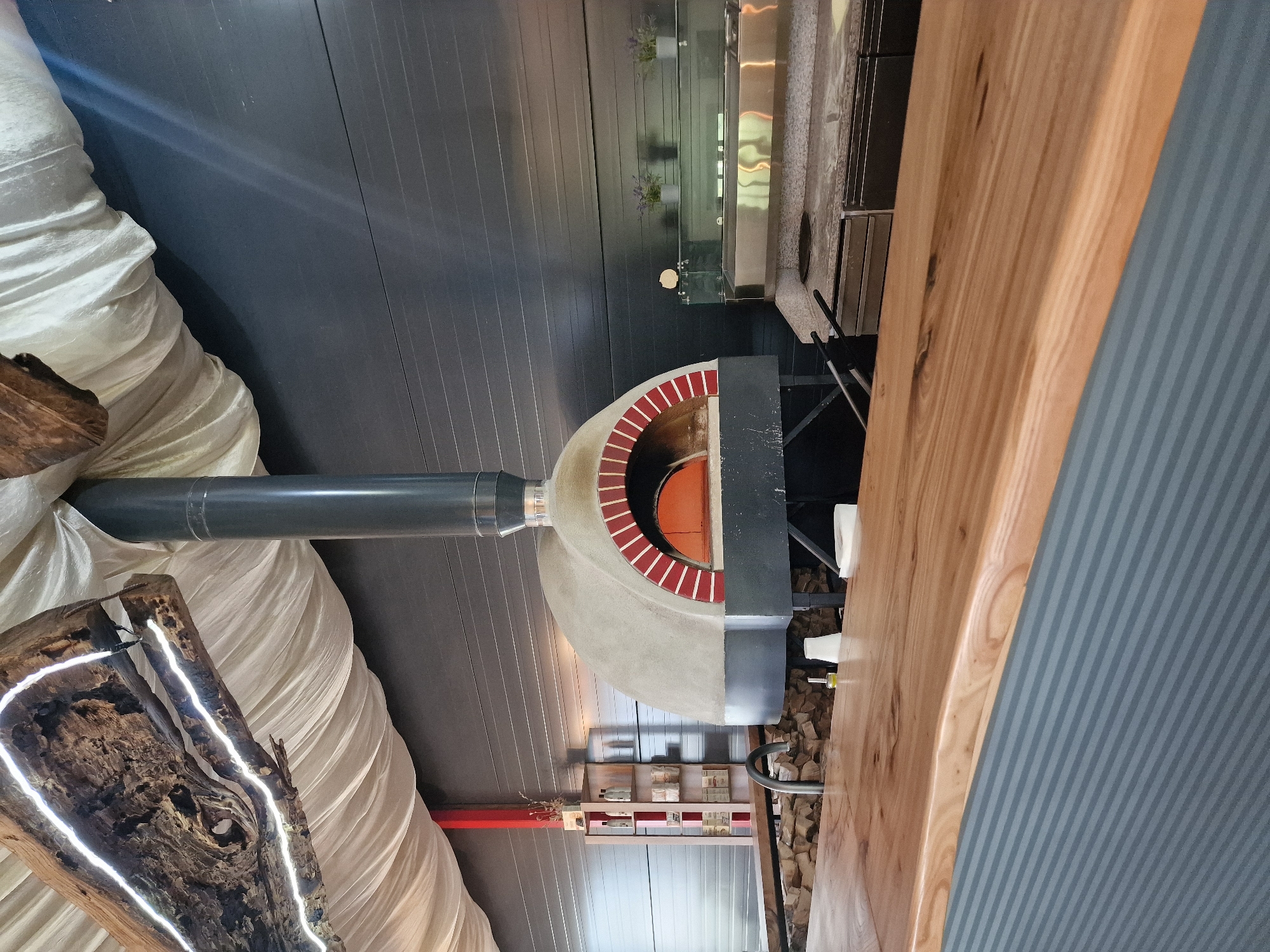 The image depicts an indoor setting with a pizza oven. The oven has a red brick archway and a gray chimney pipe extending from its top. To the left, a decorative piece of wood with embedded LED lights is partially visible. The background includes a dark gray wall with linear patterns. To the right, there is a wooden counter or table. In the distance, shelves with bottles and other items can be seen. The exact location within Aleksandrovskoye, Russia, is not discernible from the image. No people are visible in the shot.