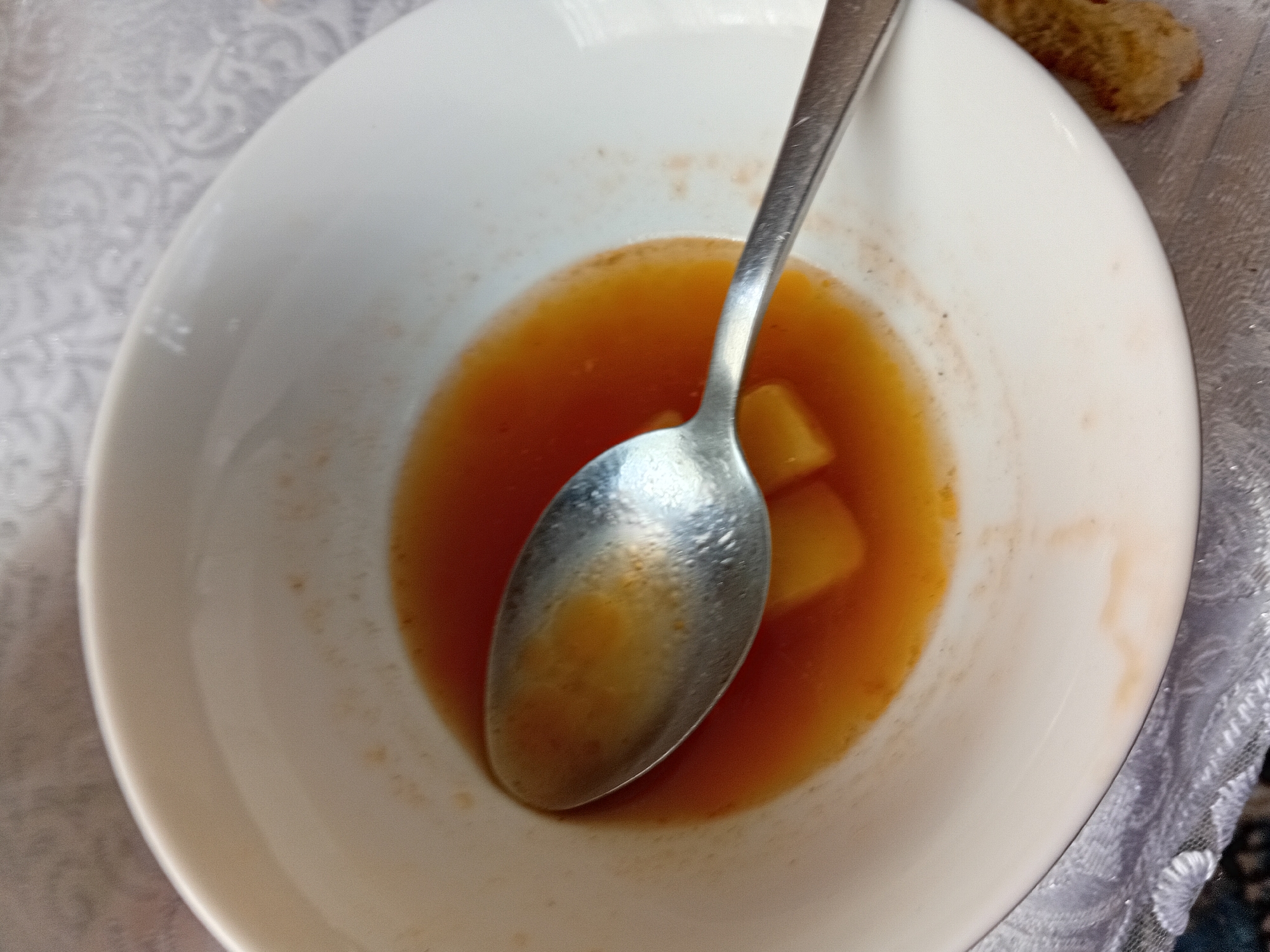 A close-up, overhead shot shows a white bowl with the remnants of a meal. A silver spoon rests in the center of the bowl, partially submerged in a small amount of orange-brown liquid. Two pieces of what appear to be cooked potato or a similar vegetable are visible beneath the spoon. The inner rim of the bowl has traces of the food. To the right of the bowl, a small piece of toasted bread is visible. The bowl sits on a white tablecloth with a subtle patterned texture. The background is out of focus, but appears to be a similar textured surface. The scene is indoors, likely a dining setting. No people are visible in the image. The location is described as Kadamjay, Kyrgyzstan, though there are no visual cues in the image to confirm this specific location.