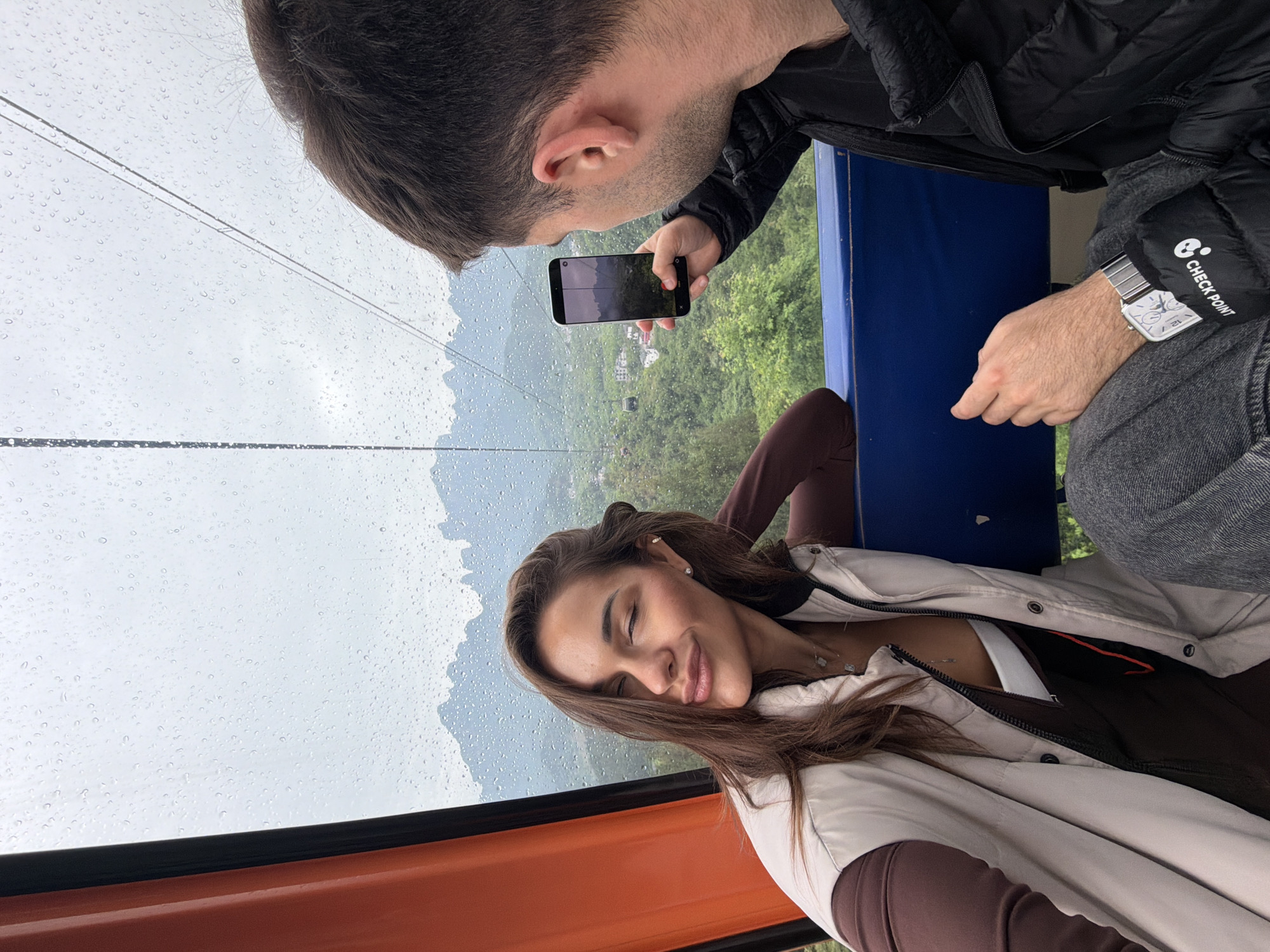A man is taking a video of a woman in a cable car on a rainy day. The cable car is suspended high above a landscape with trees and other buildings. The image was taken in the city of Zhangjiajie, China. The man is wearing a black jacket, gray pants and a watch. The woman is wearing a light-colored jacket over brown clothing. The cable car is colored orange and blue. The sky is visible through the water droplets on the glass.