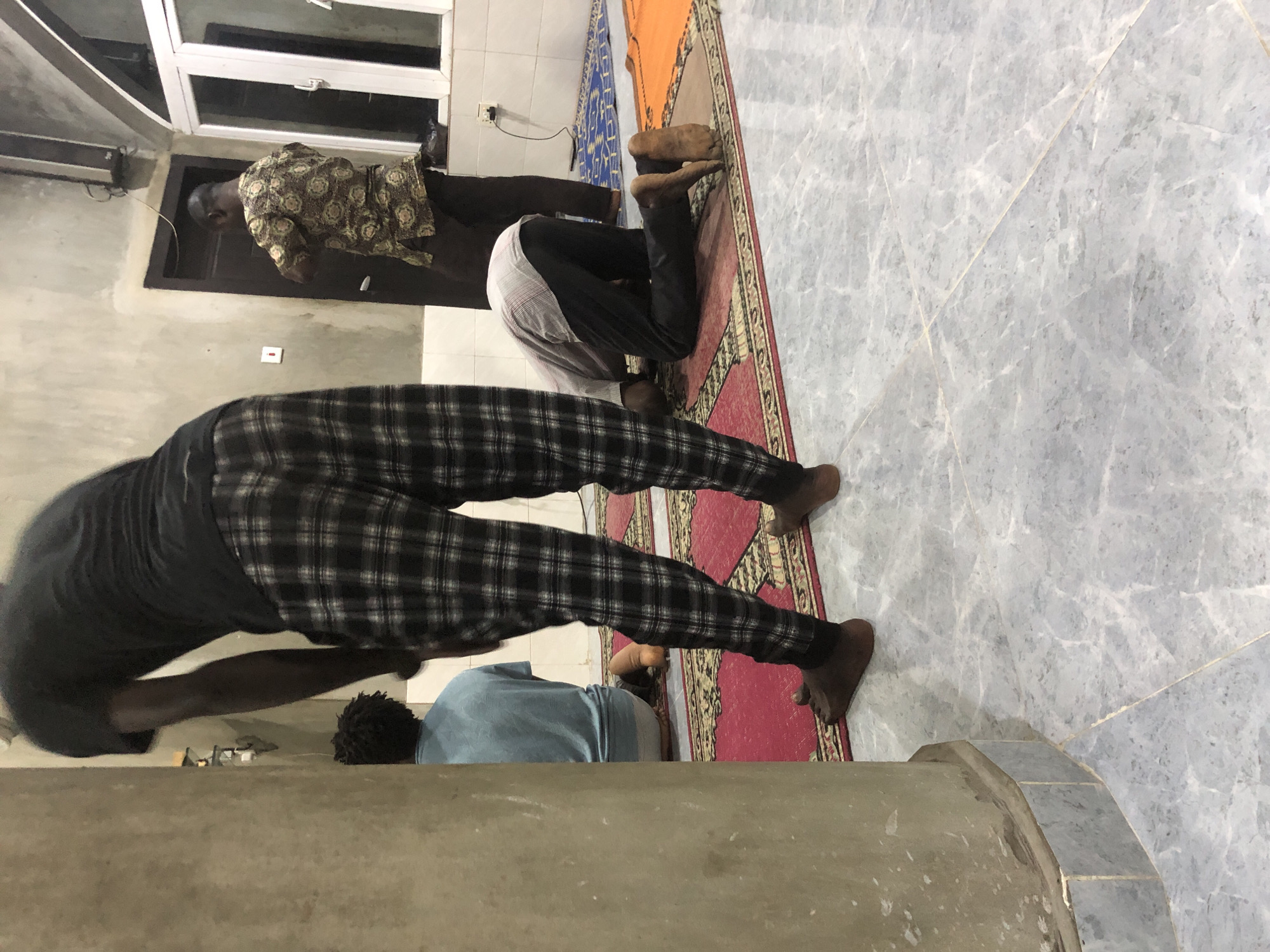 In Ado, Nigeria, inside what appears to be a prayer room, three men are engaged in prayer. The man in the foreground, on the left, stands with his back to the camera, wearing a black t-shirt and plaid pants. To his right, a man in a light blue shirt is bowed in prayer on a red rug. Further to the right, another man in a patterned shirt and dark pants is also bowed in prayer on a blue rug. The room has a light-colored, textured wall, a dark wooden door, and a large window with white frames. The floor is tiled in a grey marble pattern, and prayer rugs are laid out on top of it. A concrete pillar is visible in the left foreground. The lighting suggests an indoor environment, possibly at dusk or with artificial lighting.