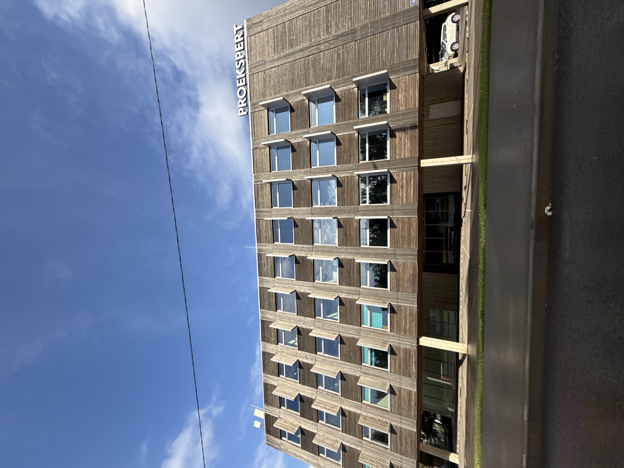 The image shows the building of Proekspert AS, a software development company located at Vana-Lõuna tänav 17 in Tallinn, Estonia. The building features a distinctive facade made of wooden planks and a series of angled window frames. The logo "PROEKSPERT" is visible at the top of the building. In front of the building, there is a wet asphalt road and a grassy area. A white car is parked in front of the building's covered entrance. The sky above is blue with some scattered clouds, and a power line runs across the upper portion of the frame.