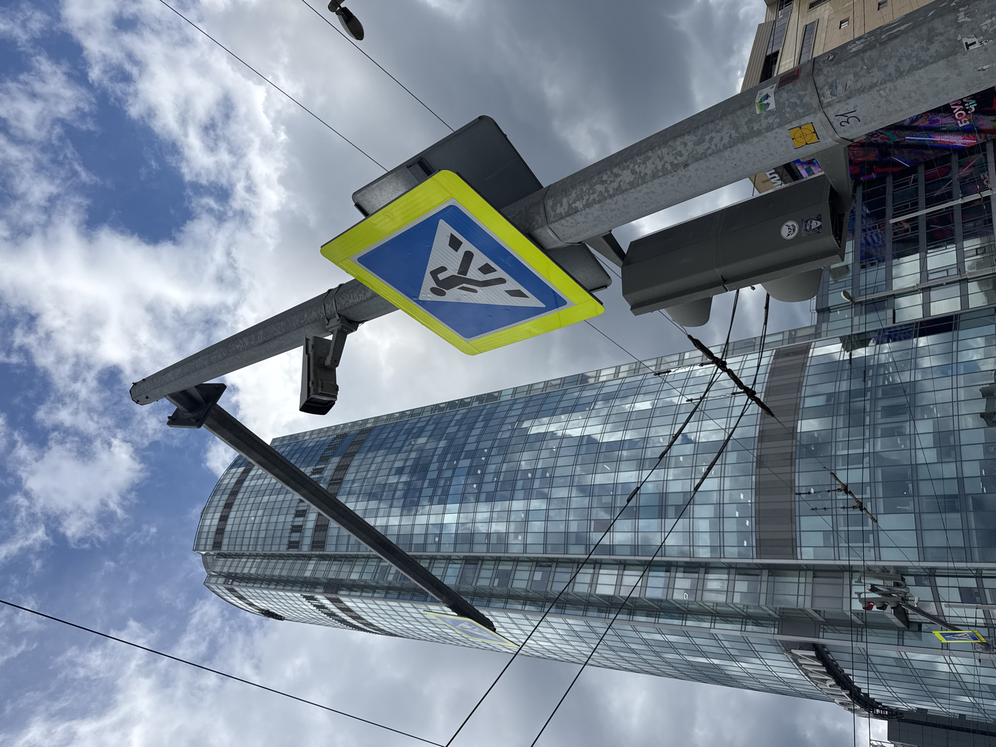 This image depicts a street scene in Yekaterinburg, Russia, featuring a modern, glass-facade skyscraper. In the foreground, a utility pole supports a pedestrian crossing sign, a traffic light, and a security camera. The pedestrian crossing sign is a yellow diamond with a blue interior, displaying a black silhouette of a person walking. The traffic light is gray and mounted horizontally. Wires, likely from an overhead power or trolleybus system, crisscross the frame, obscuring parts of the sky and the skyscraper. The sky is partly cloudy, with a mix of blue sky and gray clouds. The skyscraper, which appears to be Vysotsky Business Center based on its distinctive architecture and visible signage, dominates the background. The lower portion of the building is obscured by a street-level structure, possibly another building or commercial facade. The overall impression is an urban environment with a mix of infrastructure and modern architecture.