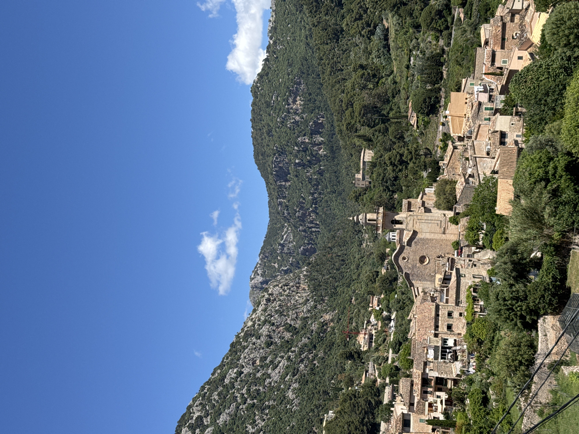The image shows an aerial view of the town of Valldemossa in Spain. The scene depicts a mountain landscape with lush green vegetation covering the hillsides. The town is built on the slopes of the mountain, with buildings densely clustered together. The architecture of the buildings features traditional Spanish style, with terracotta roofs and stone walls. A prominent church with a round window is visible in the center of the town. The sky is a clear blue with a few scattered clouds. The perspective suggests the photo was taken from an elevated position, possibly a cable car or another high point in the area.