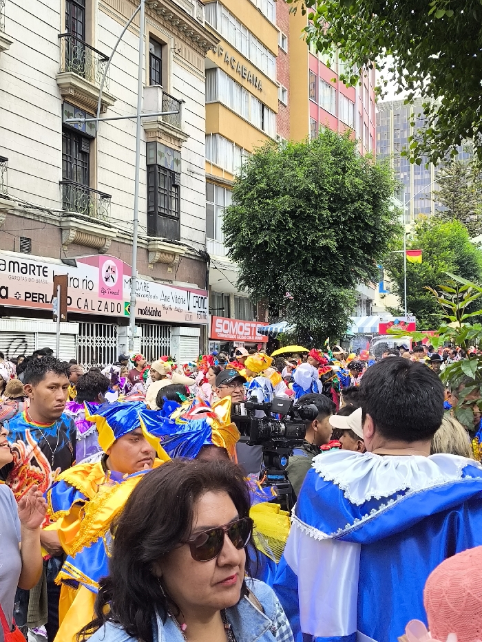 A street scene features a dense crowd of people, many dressed in vibrant blue and yellow costumes. A woman with sunglasses and a denim jacket stands in the foreground, looking to the side. A professional video camera is visible in the middle of the crowd. Buildings line the street, including one with the sign "PACAEBANA". Storefront signs and a Bolivian flag add to the scene, suggesting a festive or parade-like atmosphere.