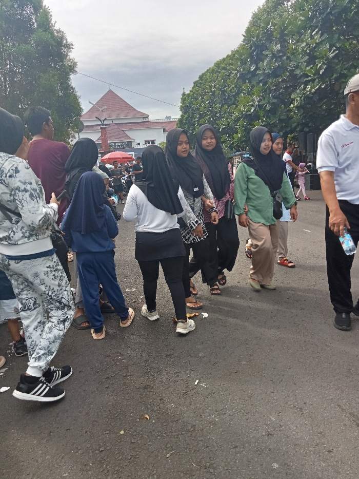This daytime outdoor scene in Singaparna, Indonesia, captures a diverse group of people, predominantly women and girls wearing hijabs, moving along an asphalt road under an overcast sky. People are dressed in casual attire, including various long-sleeved tops, pants, leggings, and tracksuits, with footwear ranging from sneakers (some with Adidas branding) to sandals and flip-flops.
In the foreground, a young girl in a blue hijab and tracksuit walks with her back to the camera, near a woman in a white top and black leggings. To their right, several women in hijabs are walking together, some looking towards the camera, others engaged with each other. On the far right, a man in a white polo shirt, partially visible, carries a plastic water bottle.
In the mid-ground, motorcycles and a red canopy suggest further activity. The background features a prominent building with a distinctive reddish-brown tiled roof, flanked by lush green trees. Partially visible text on the building identifies it as "KANTOR BUPATI TASIKMALAYA," indicating the Regent's Office of Tasikmalaya. The general movement and casual dress suggest a public gathering, a walk, or an event. People appear relaxed and engaged with their surroundings.