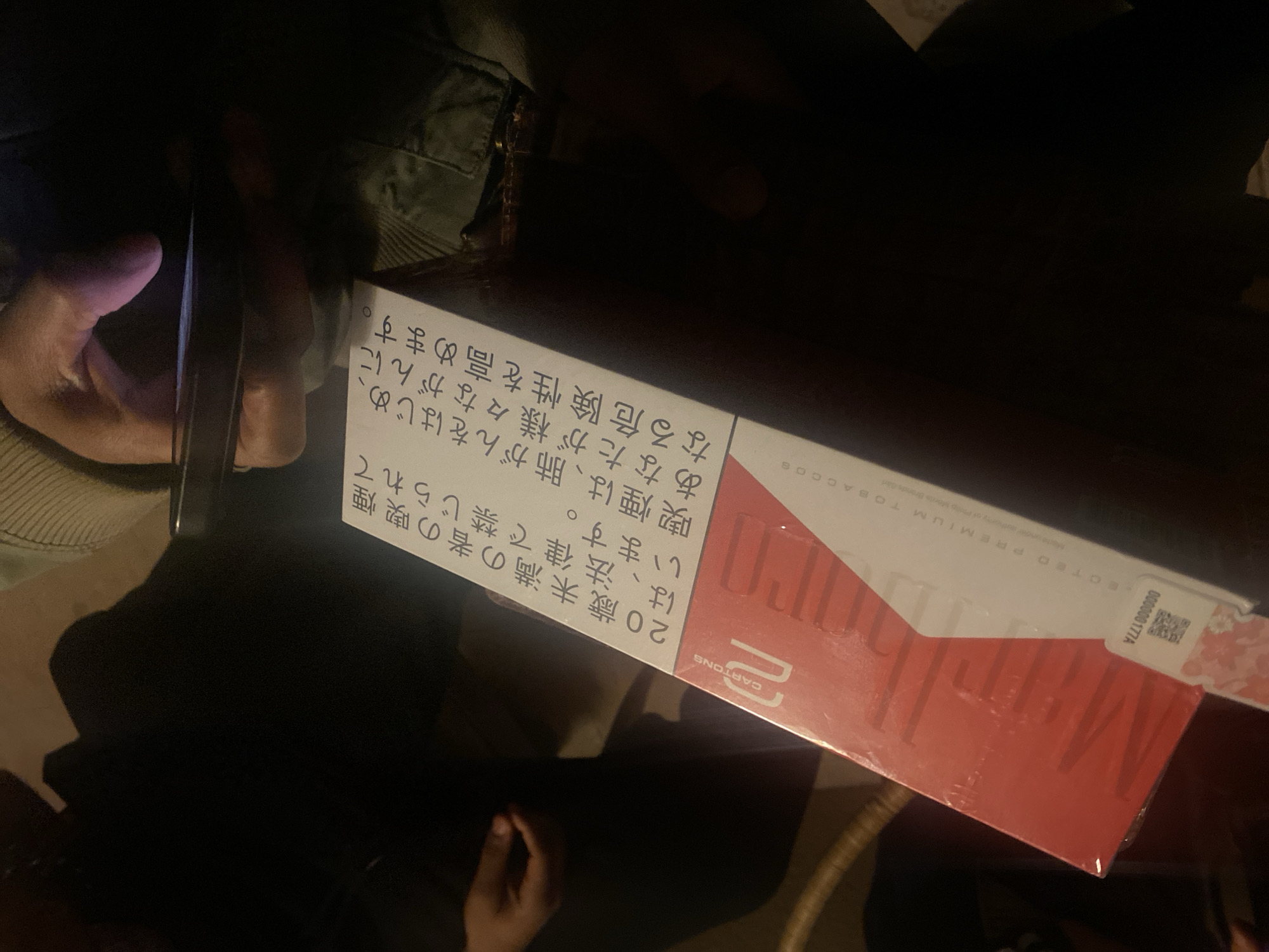A person is holding a smartphone pointed at a pack of cigarettes in a poorly lit environment. The cigarette pack is red and white, and has Japanese text on it. The person's skin appears to be dark. A portion of another person's clothing is visible in the upper-left. The location appears to be indoors. There is no explicit information in the image to confirm it is located in the city of Montreuil, France.