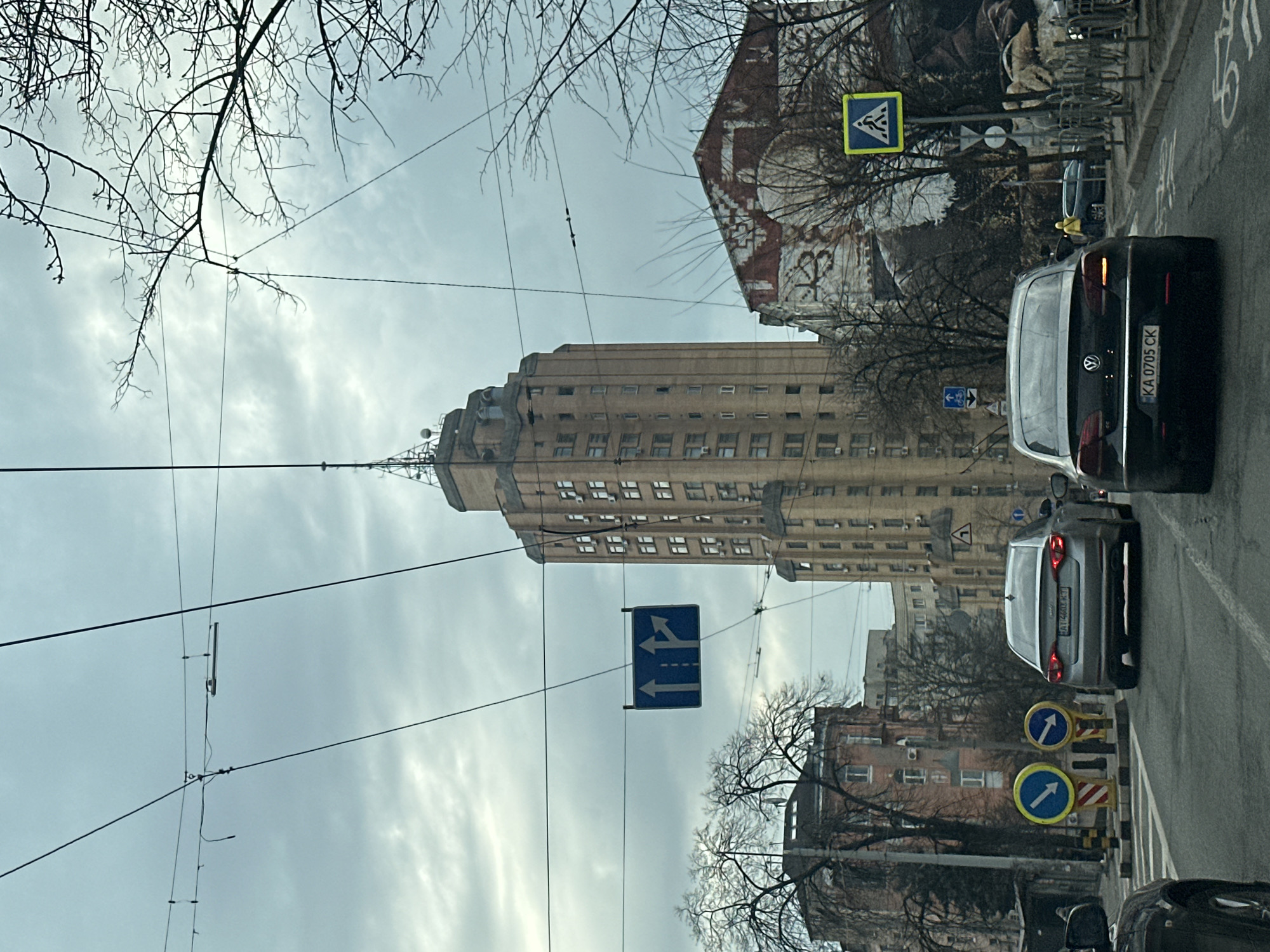 The image depicts a street scene in Kyiv, Ukraine, dominated by the tall, multi-story building located in Kyiv, Ukraine at Kreschatyk Street. The building has a distinctive tiered shape, with rows of windows and a spire-like structure on top. The sky is overcast. Overhead power lines are strung across the sky. The street is lined with buildings, including older buildings with decorative elements, buildings with graffiti, and several street signs. Vehicles are present, including several cars, one of which has the license plate "KA 0705 CK". Other road signs and construction barriers are present on the right side of the street. There are bare trees lining the street.