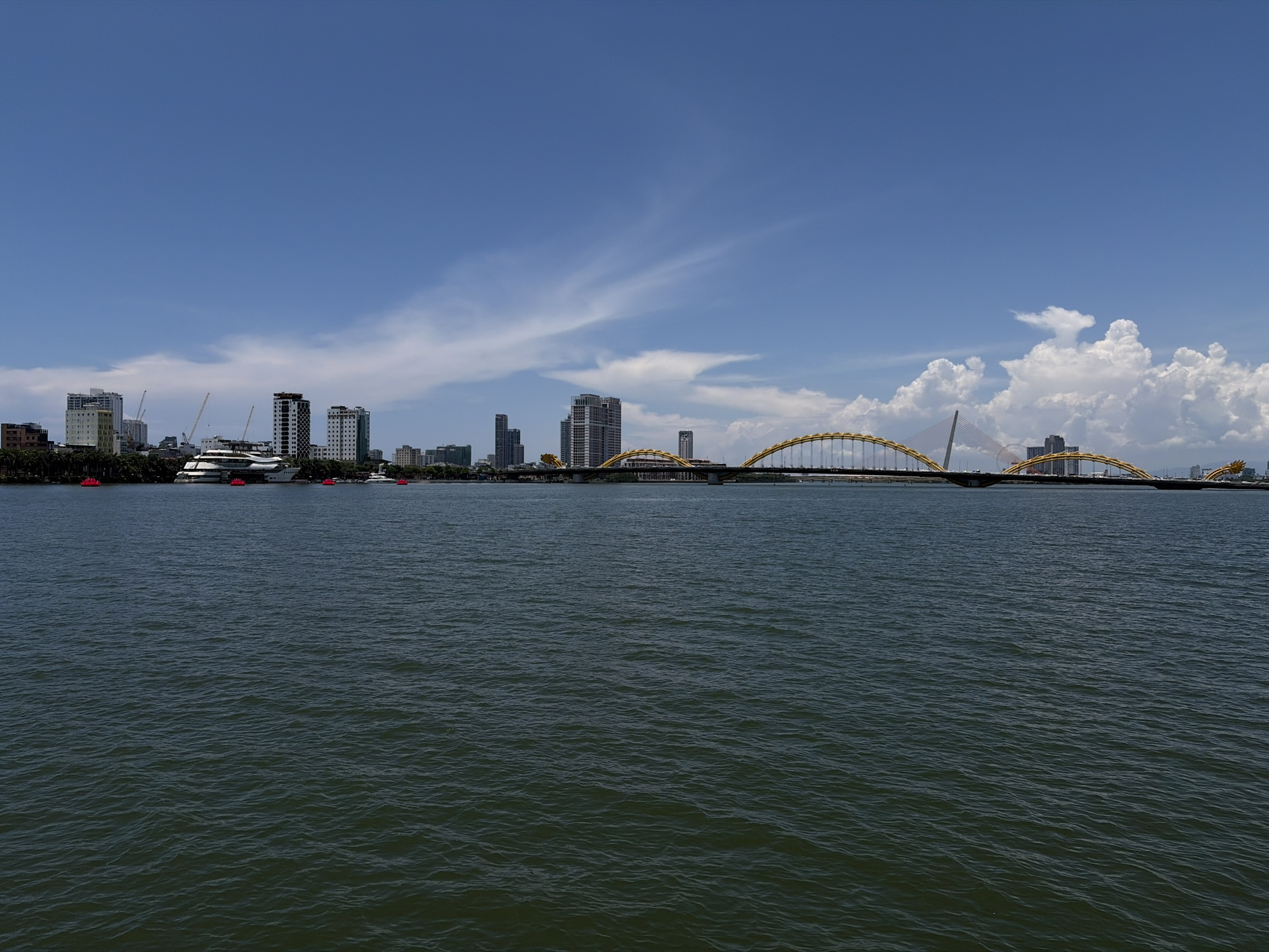 This wide shot captures a cityscape and the Dragon Bridge spanning the Han River in Đà Nẵng, Vietnam. The water occupies approximately the lower half of the frame, showing gentle ripples. Above the water, the city skyline includes several tall buildings and construction cranes. The Dragon Bridge, with its distinctive golden dragon head, arches across the river. The sky is blue with scattered clouds, suggesting a clear, sunny day. A white yacht is docked closer to the left side of the city skyline. A few small boats, marked with red buoys, are visible between the yacht and the city.