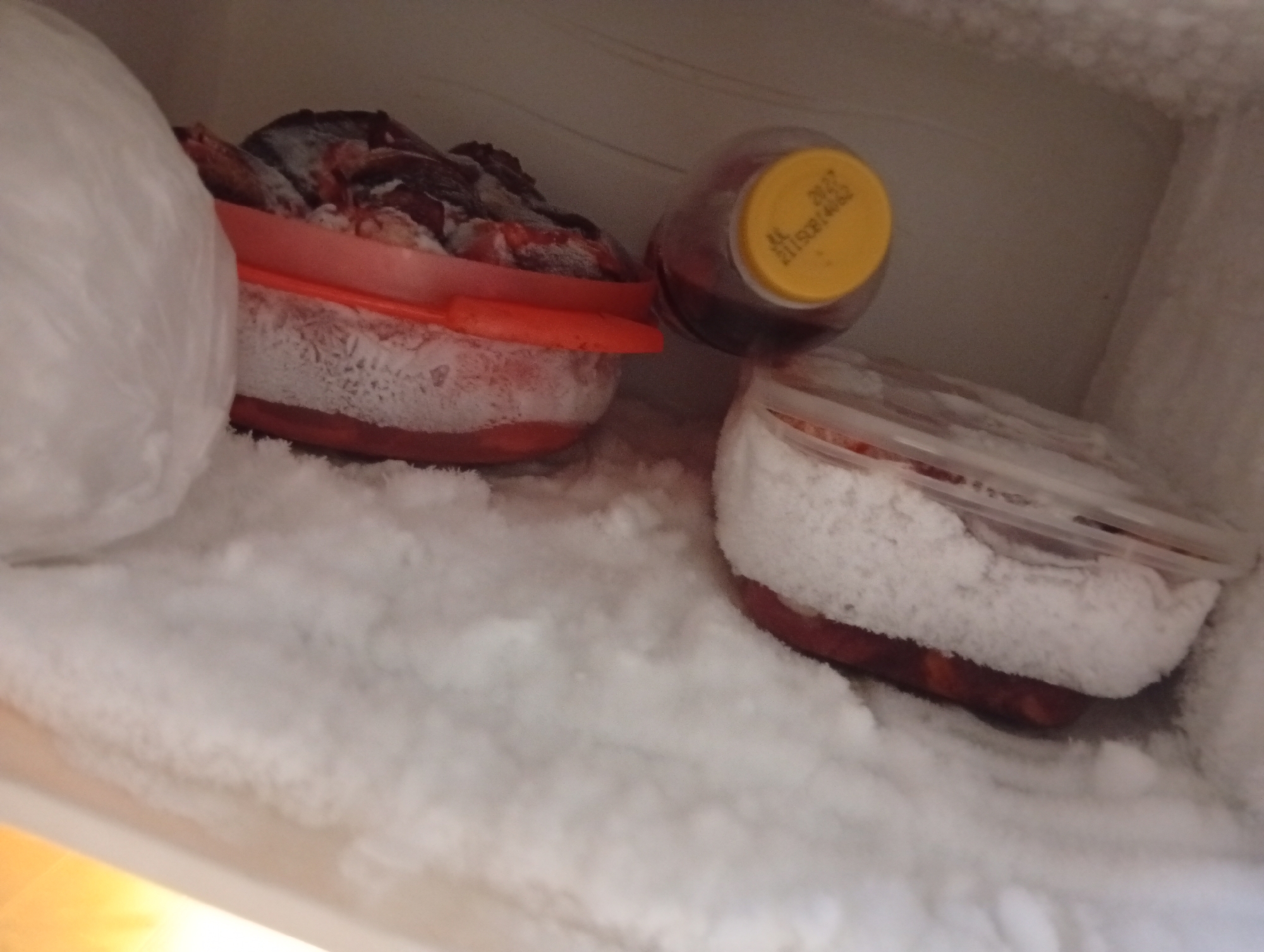 A close-up, eye-level, indoor shot displays a freezer compartment with a significant accumulation of frost. To the left, a large, white, rounded object, possibly a bag of frozen food, is partially visible. In the center, a red plastic container filled with dark-colored food, likely meat, is covered in frost. Next to it, a clear glass jar with a yellow lid contains a dark liquid. The interior of the freezer is heavily frosted, obscuring some details and creating a textured, white surface. The lighting is dim, typical of an interior freezer environment, with a warm light source visible at the bottom left of the frame. The scene is static and shows no signs of human activity or interaction with the objects. The location is described as Kualu, Indonesia, but there are no visual cues in the image to confirm this geographical context.