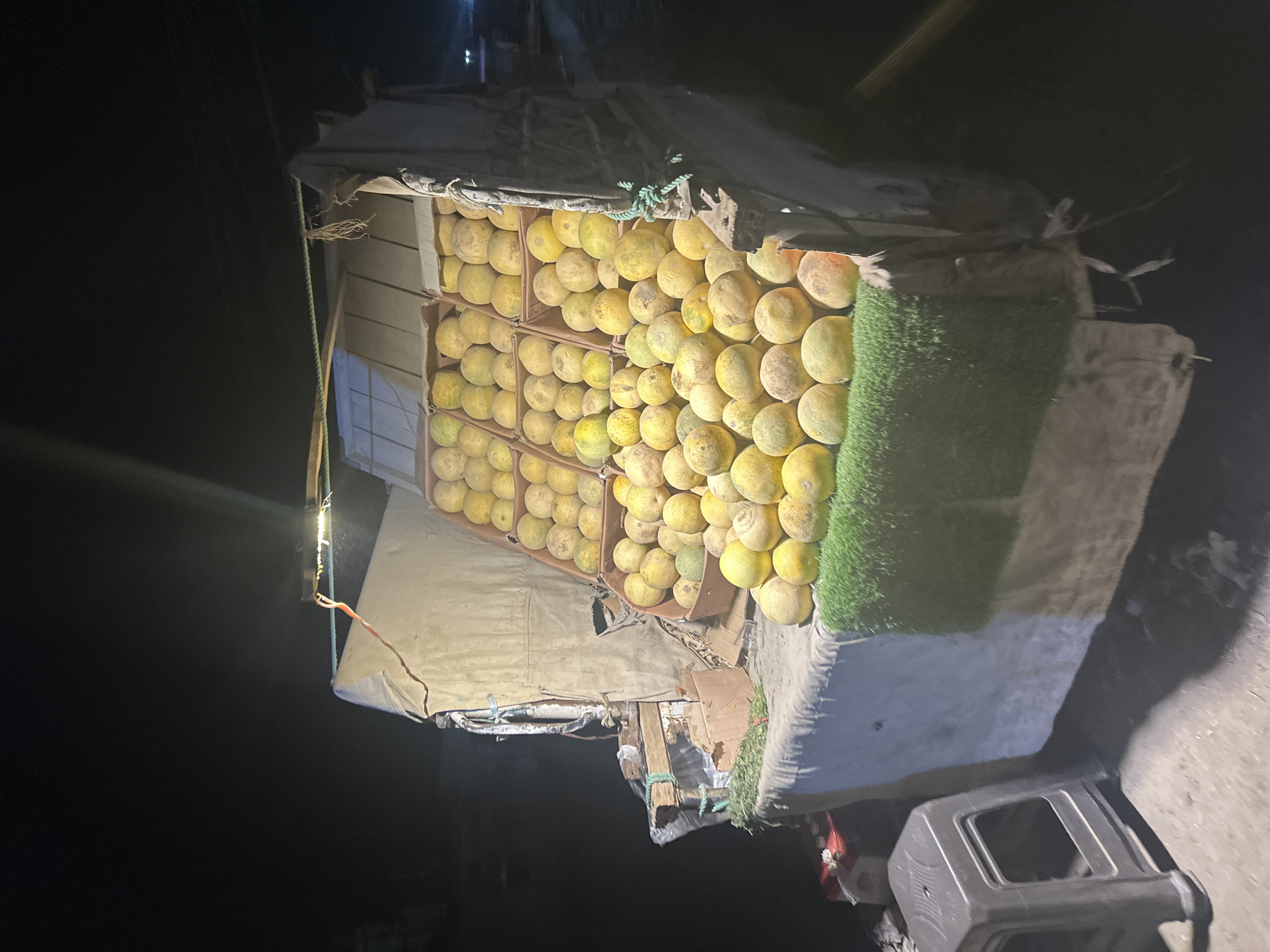 A makeshift market stall in Quetta City Tehsil, Pakistan, displays stacks of ripe yellow melons in cardboard boxes under a canvas roof. A section of artificial green turf decorates the stall's edge. The scene is illuminated by a bright light source in the dark night environment, with a plastic stool visible at the base.