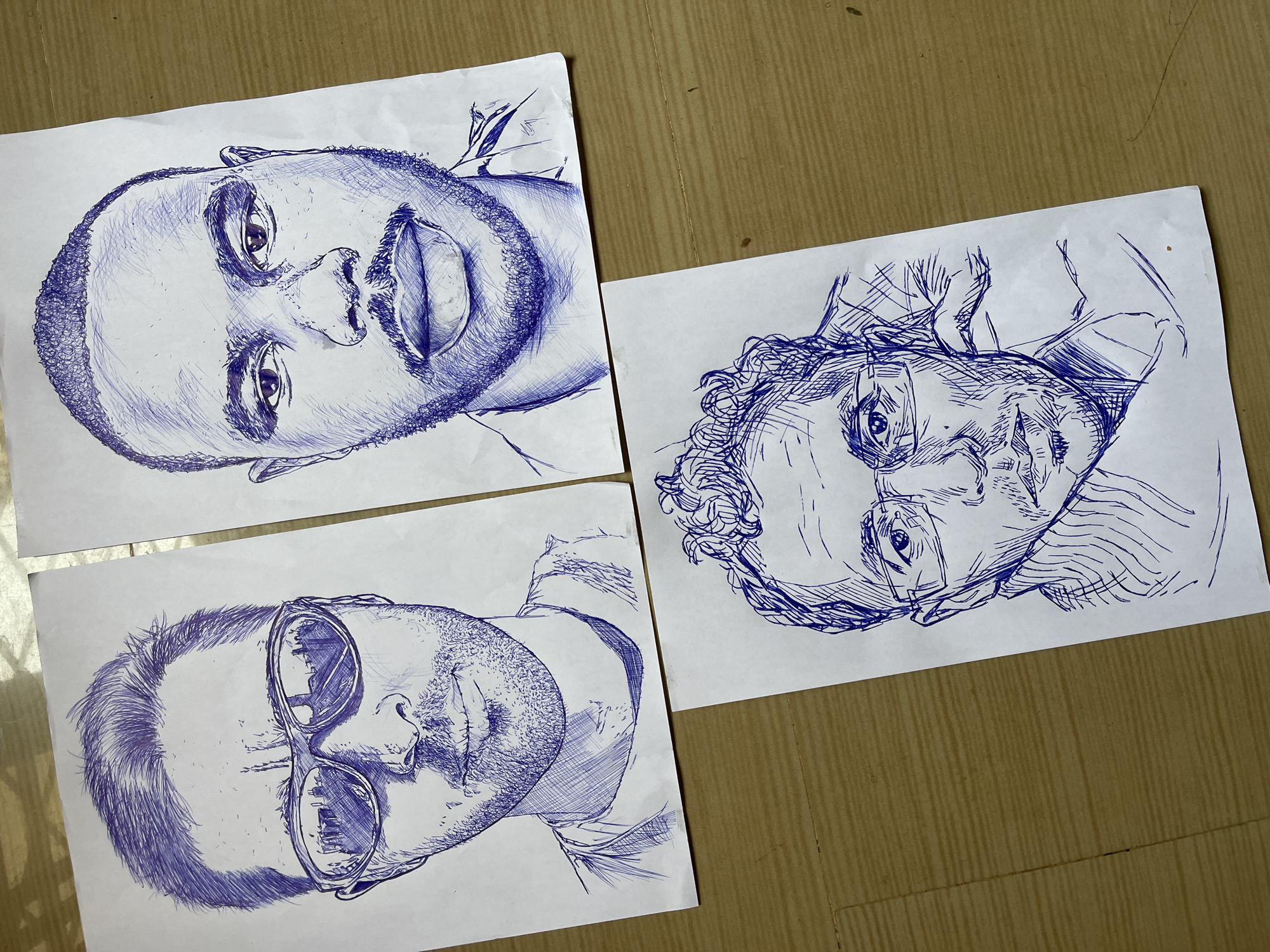 Three pen-and-ink portraits of men are laid out on a light brown, textured surface. The portraits are drawn on white paper, with the ink appearing to be blue. The surface resembles a wooden floor or table with a distinct grain pattern.
The top-left portrait depicts a man with short hair, wearing sunglasses. The lenses of the sunglasses reflect an urban landscape, possibly buildings and a road. The man has a stubbled beard and is looking directly at the viewer.
The top-right portrait is of a Black man with short, textured hair and a beard. He is also looking directly at the viewer, with a neutral expression. His lips are slightly parted.
The bottom portrait features a man with curly hair and glasses, resting his chin on his hand. He is looking off to the side with a contemplative expression. His beard is visible, and he is wearing a collared shirt.
The image does not provide any specific information about the location being Jos, Nigeria. The setting is generic and appears to be an indoor surface.