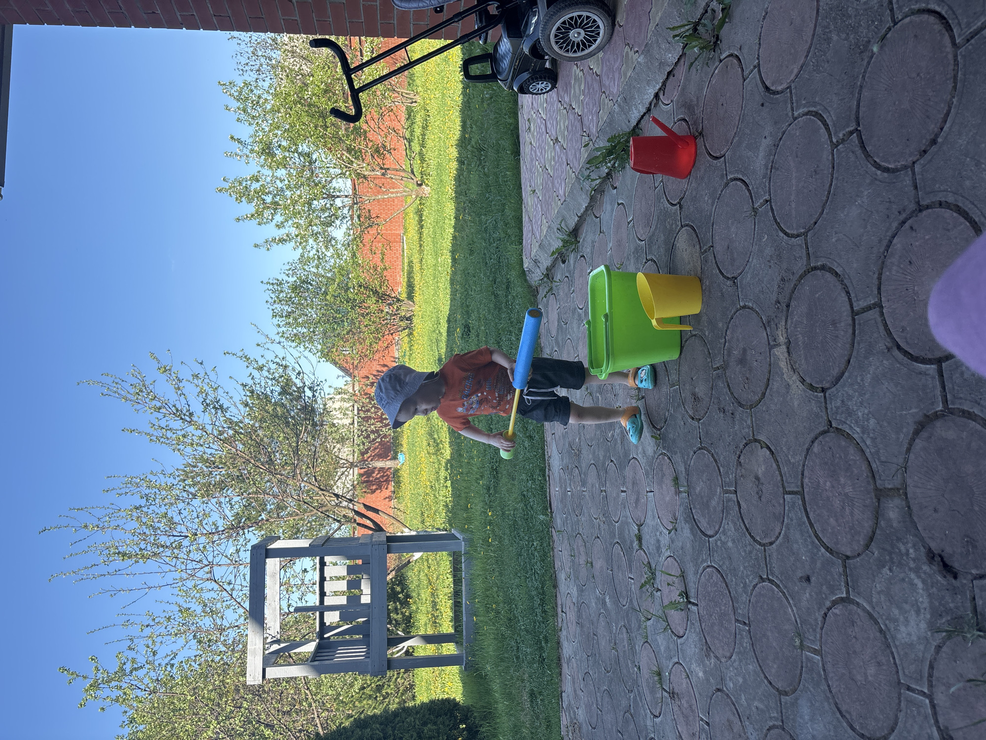 A child stands on a brick-laid patio in Ферма, Russia. He wears a gray hat, an orange shirt, and dark shorts. He holds a blue toy bat and a yellow ball. A green bucket and yellow cup are next to him. A red watering can is nearby. A black lawnmower sits on the patio. Behind him is a grassy yard with a brick wall, a tree, and a gray wooden structure. The sky is clear blue.