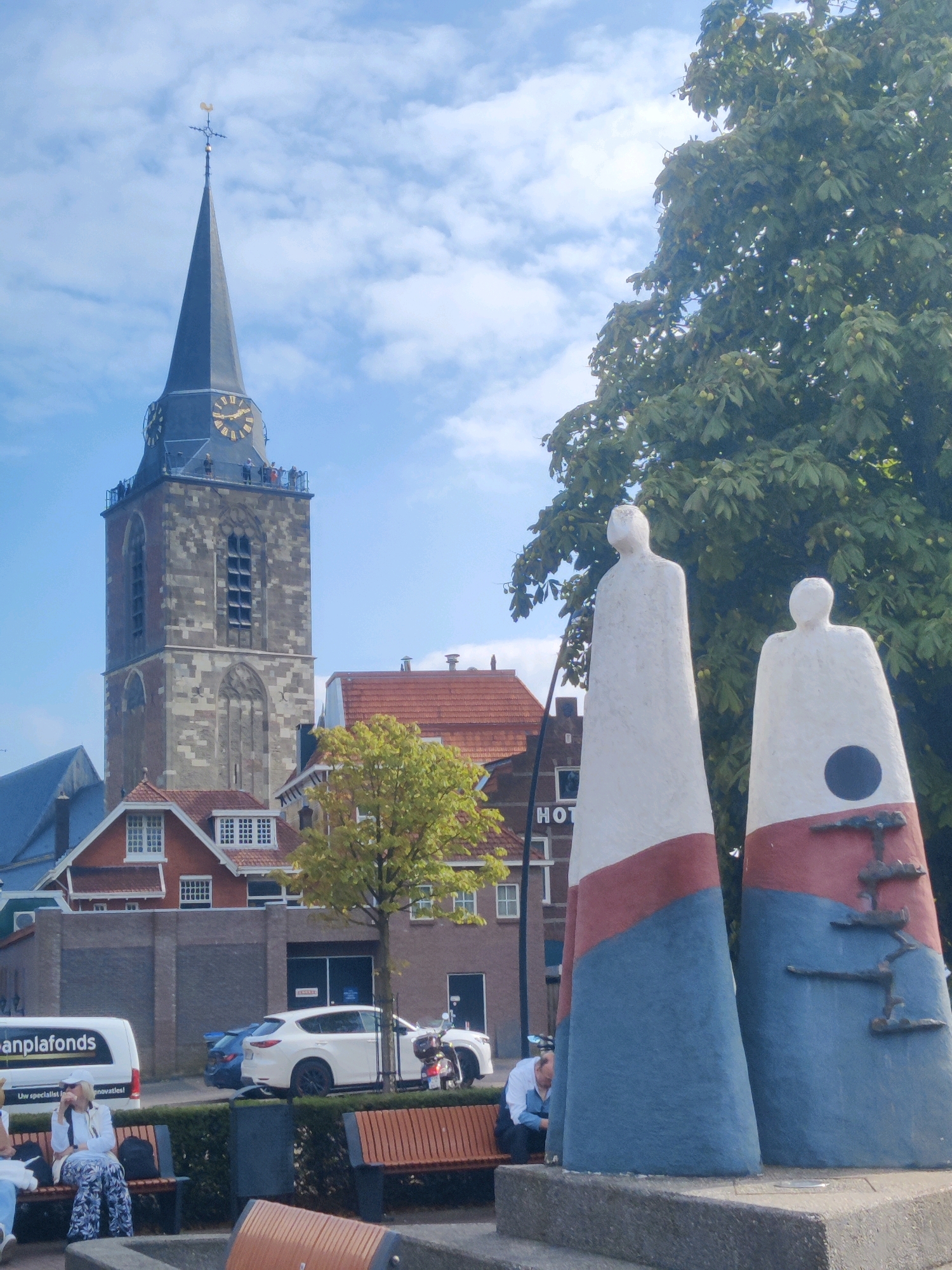 The image depicts a street scene in the city of Winterswijk, Netherlands. In the foreground, two tall, abstract sculptures with red and blue bands stand prominently. To the left of the sculptures, a woman is seated on a park bench, dressed in a white jacket and patterned pants. To the right, a man is also seated on a bench, leaning forward. Behind the sculptures and benches, a white van with the text "anp" and "plfonds" is parked. Further back, several buildings are visible, including a prominent church tower with a clock face. A white car and a motorcycle are parked on the street. A large tree with green leaves and some seed pods is situated to the right of the sculptures. The sky is blue with scattered clouds.