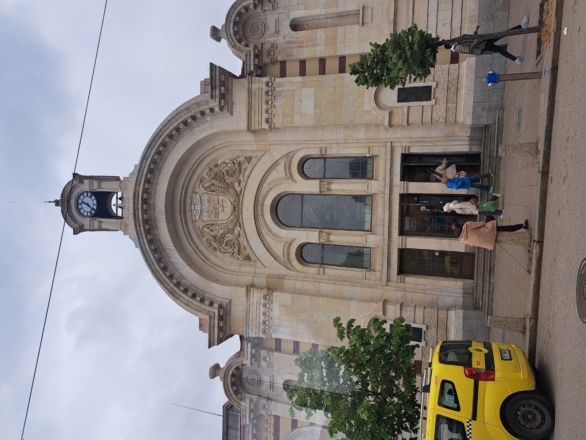 The image depicts the facade of the Saint Sofia Church in Sofia, Bulgaria. It's a beige stone building with a large arched entrance, ornate carvings, and a clock tower. Several people are seen entering or exiting the church. A yellow taxi is parked on the street alongside the building. The sky is overcast.