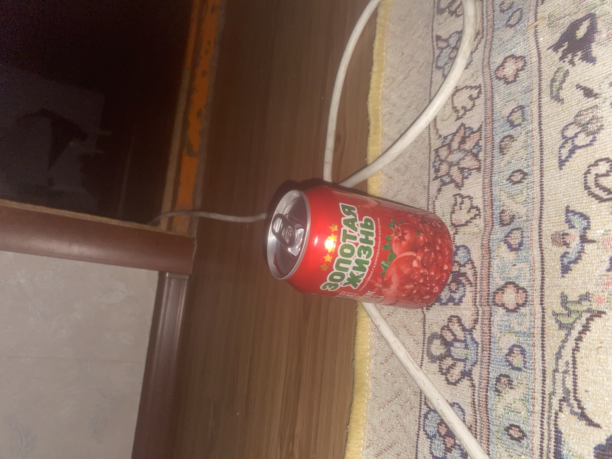 A red soda can, branded with Russian text and an image of pomegranates, sits on a patterned rug. A white cable runs across the rug and behind the can. The background consists of a wooden floor and a dark, blurry area that could be furniture or a doorway. The scene appears to be indoors, possibly in a home. There is no indication of the location being in Ойтал ауылдық округі, Kazakhstan.