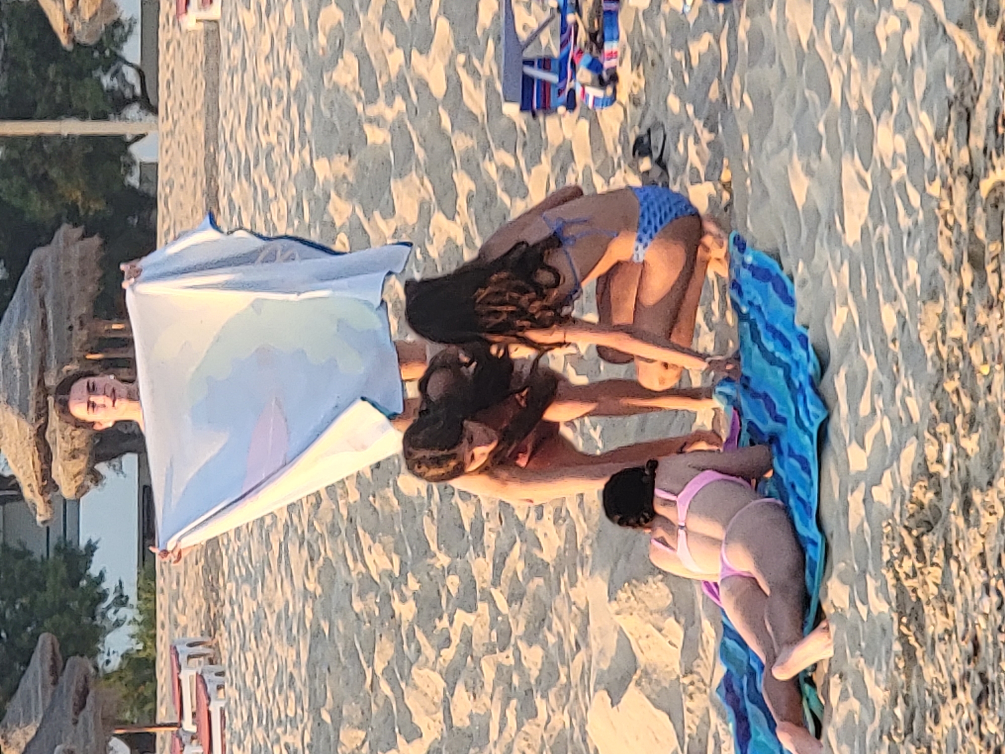 The image captures a scene on the sandy beach of Quarteira, Portugal. In the foreground, three young females are present. One, positioned on a blue and green striped towel, is lying on her stomach, facing away from the camera, wearing a pink bikini. To her right and slightly behind her, another female is crouched down, wearing a blue bikini with a patterned design, looking towards the ground. A third female is standing between them, bending forward and holding up a large white and blue patterned beach towel. Her face is visible as she looks forward. In the background, beach umbrellas with thatched roofs and red-and-white sun loungers are visible. The sand is textured with footprints and shadows.