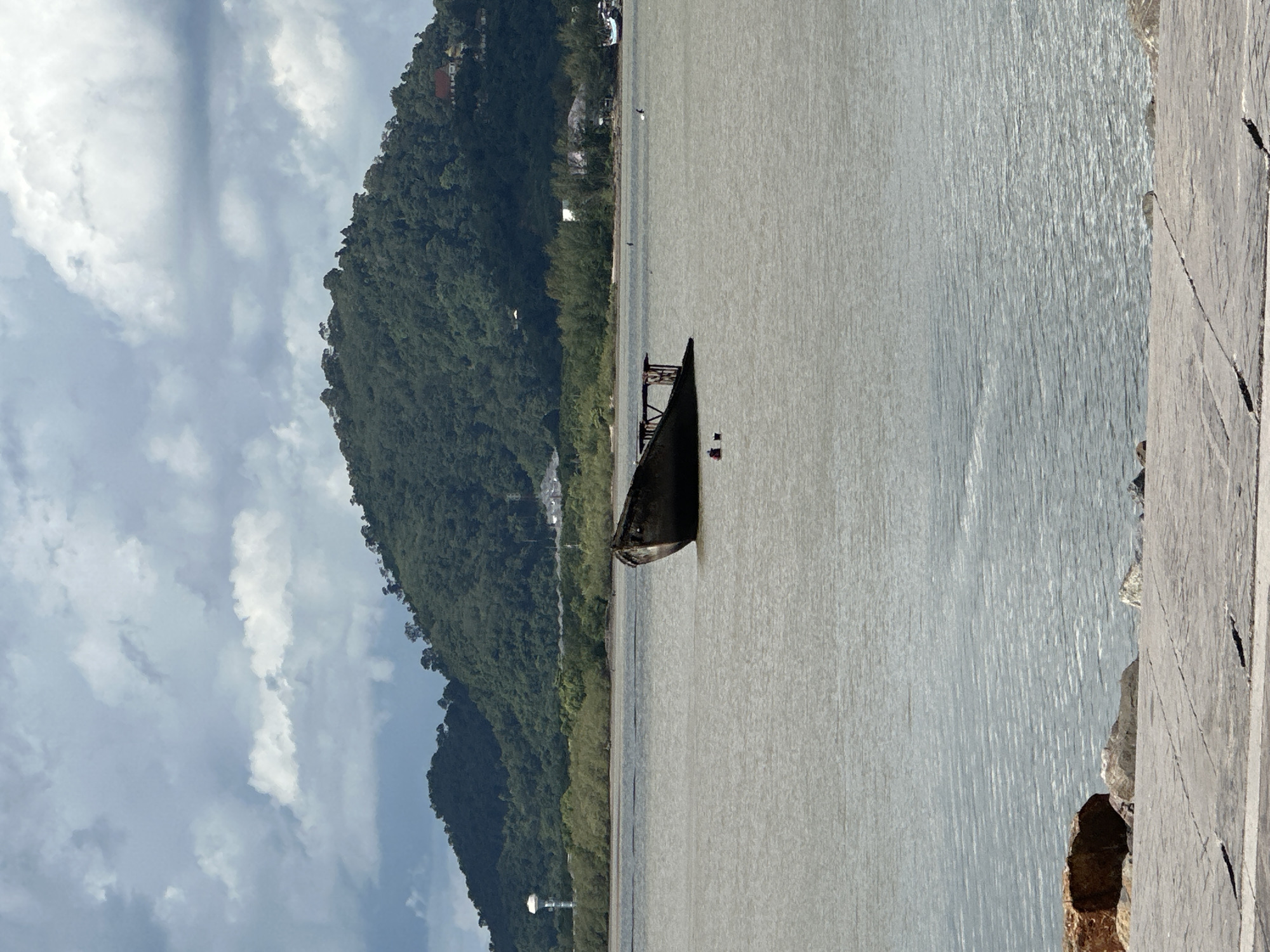 An abandoned, rusting shipwreck sits partially submerged in a wide, murky body of water, likely the Andaman Sea near Phuket City Municipality, Thailand. The vessel, with its skeletal superstructure exposed, appears to be listing heavily to starboard. In the background, lush, green hills rise steeply, their slopes densely covered with trees. A water tower and some buildings are visible on the slopes, indicating a populated area. The sky is overcast with dramatic, dark clouds, suggesting the possibility of rain. The foreground consists of a paved walkway or pier with large rocks and stones scattered along its edge. The overall scene conveys a sense of decay and abandonment against a backdrop of natural beauty.