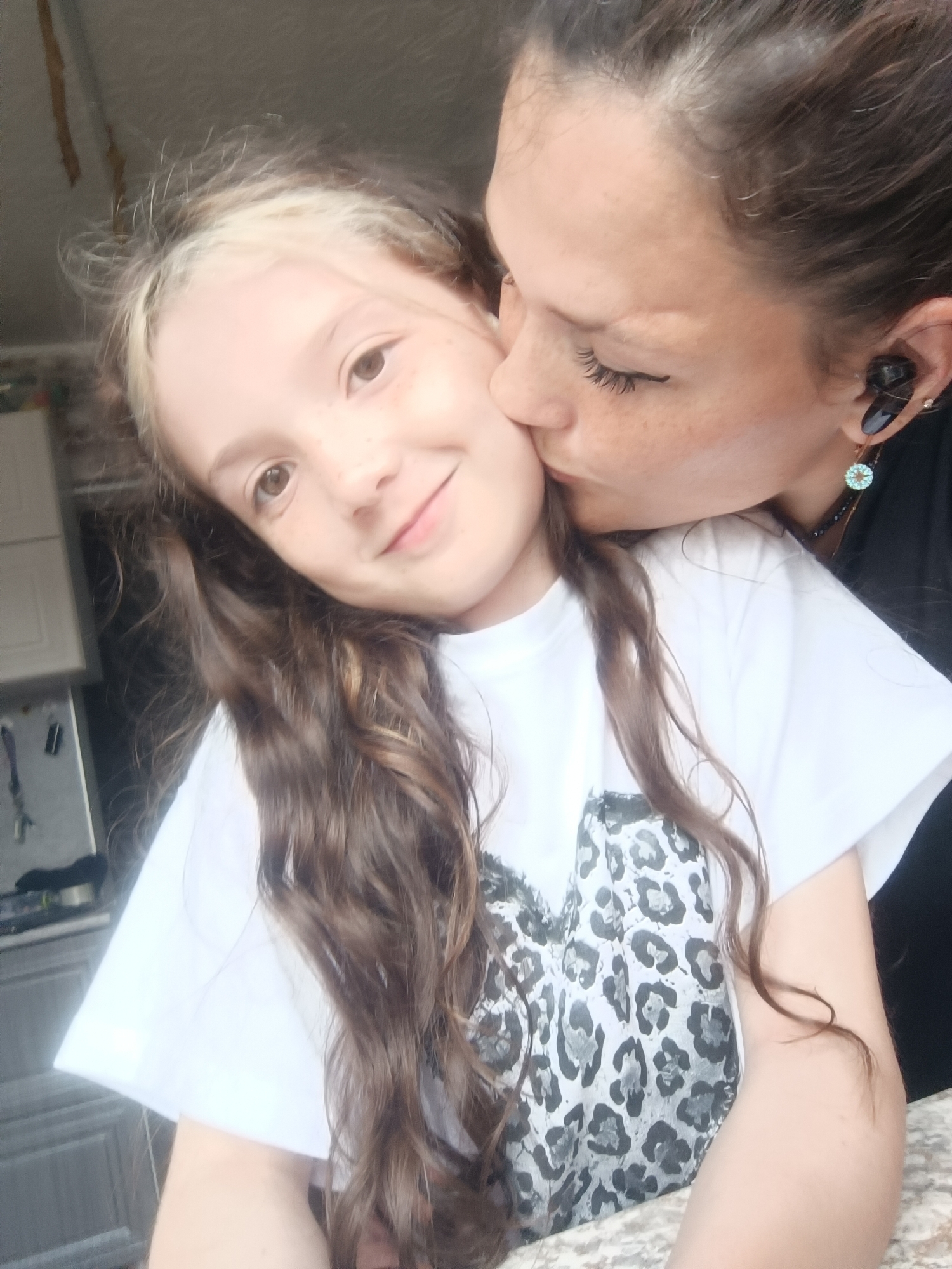 In this medium shot, a woman is kissing a young girl on the cheek. Both individuals are in a close-up shot, with the girl positioned slightly in front of the woman. The girl has long, wavy brown hair with streaks of blonde and is wearing a white t-shirt with a leopard print design. Her eyes are open and she is smiling. The woman has brown hair and is wearing black. She has on earbuds in her right ear and earrings. The background appears to be indoors, possibly a kitchen, with cabinets and a countertop visible. The lighting suggests it might be daytime, but the weather is not discernible. The overall emotion conveyed is affection and tenderness. There is no visible text in the image.