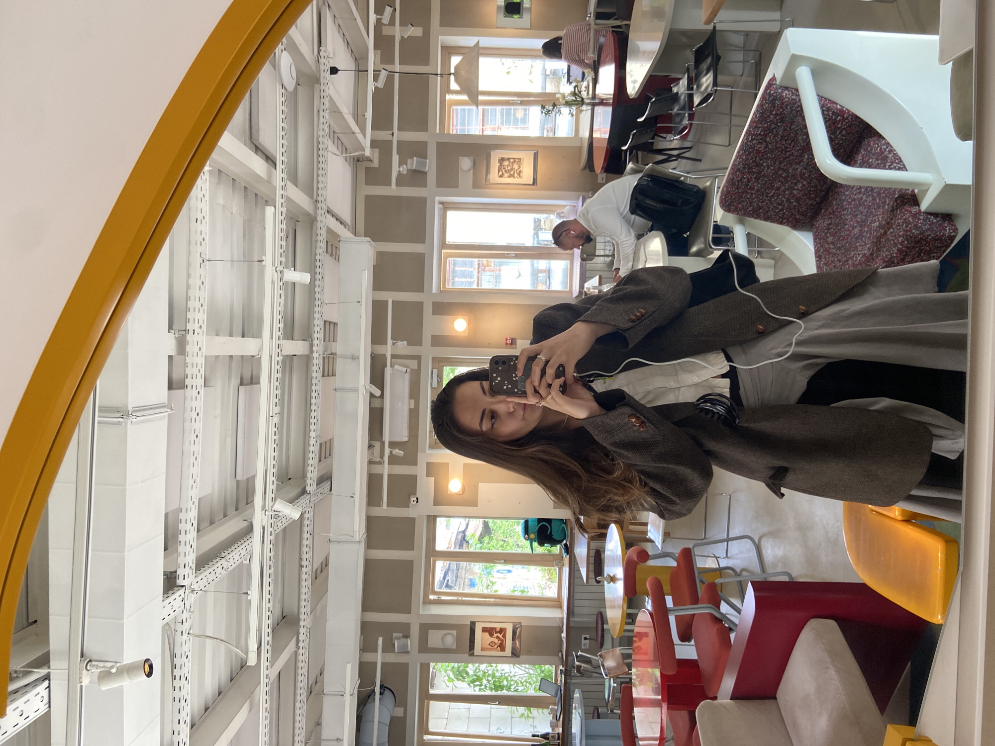 A woman is taking a selfie in a mirror. She is wearing a brown blazer and holding a smartphone. The mirror's frame is partially visible, colored in a yellow-orange hue. The location appears to be a café or restaurant. The interior features a high ceiling with exposed ductwork and hanging lights, painted in white. Windows are spaced throughout the room, providing natural light. Tables and chairs, including some upholstered seating in red and beige tones, are visible. Other people are present in the background, including individuals seated at tables. This image was taken in Moscow, Russia.