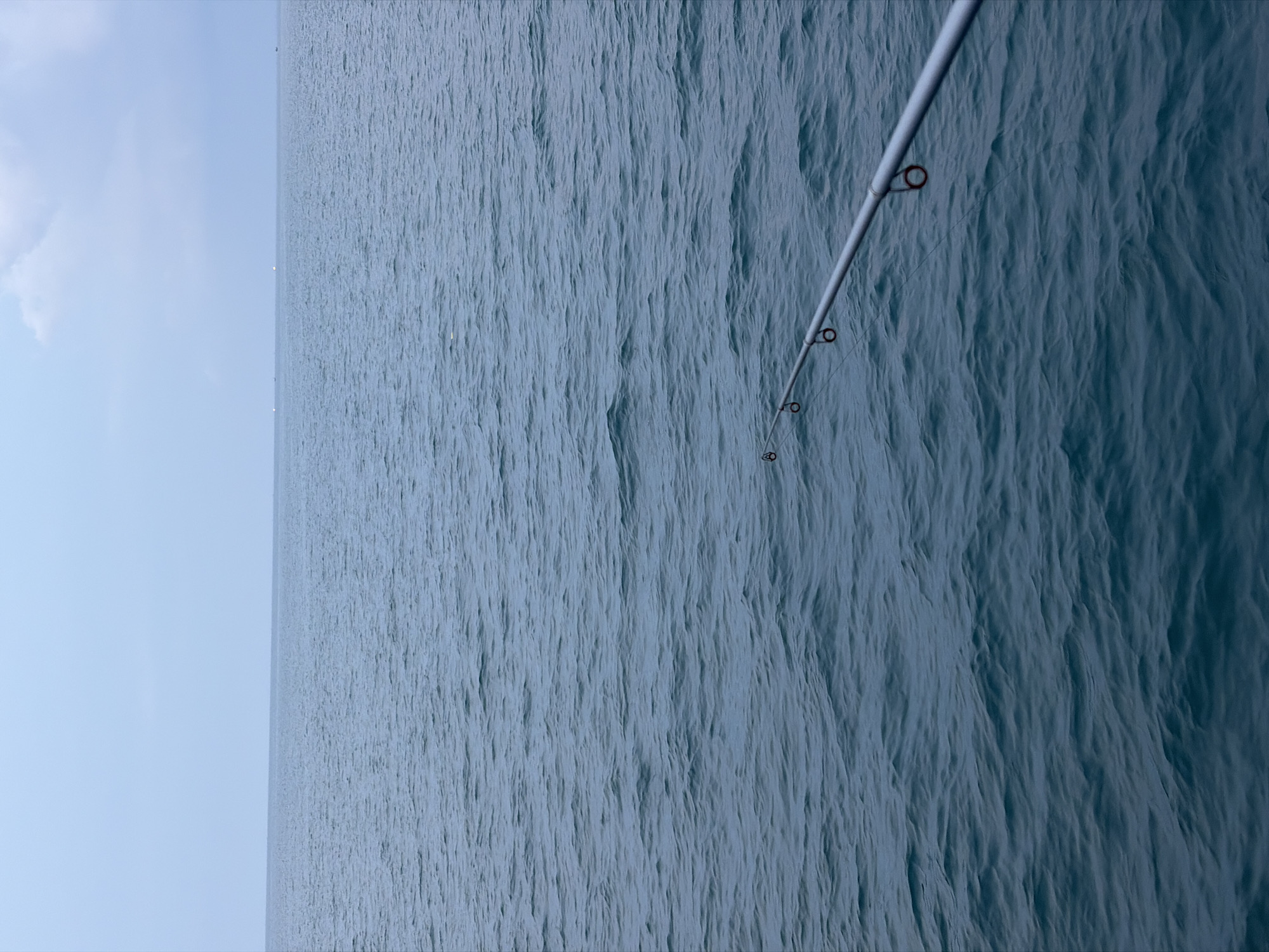 The image depicts a serene view of the ocean, likely from a fishing boat or pier. The water is a deep blue-green, with gentle ripples on the surface indicating a calm day. In the foreground, a fishing rod extends diagonally from the bottom right corner towards the left, with its line disappearing into the water. The rod is silver and has several guide rings. The horizon line is distinct, separating the water from a pale, overcast sky. A few faint lights or distant vessels are visible on the horizon. The scene suggests a quiet moment of fishing or enjoying the coastal environment. This image could be from the waters off Lhokseumawe, Indonesia, a city known for its coastal location and fishing activities.