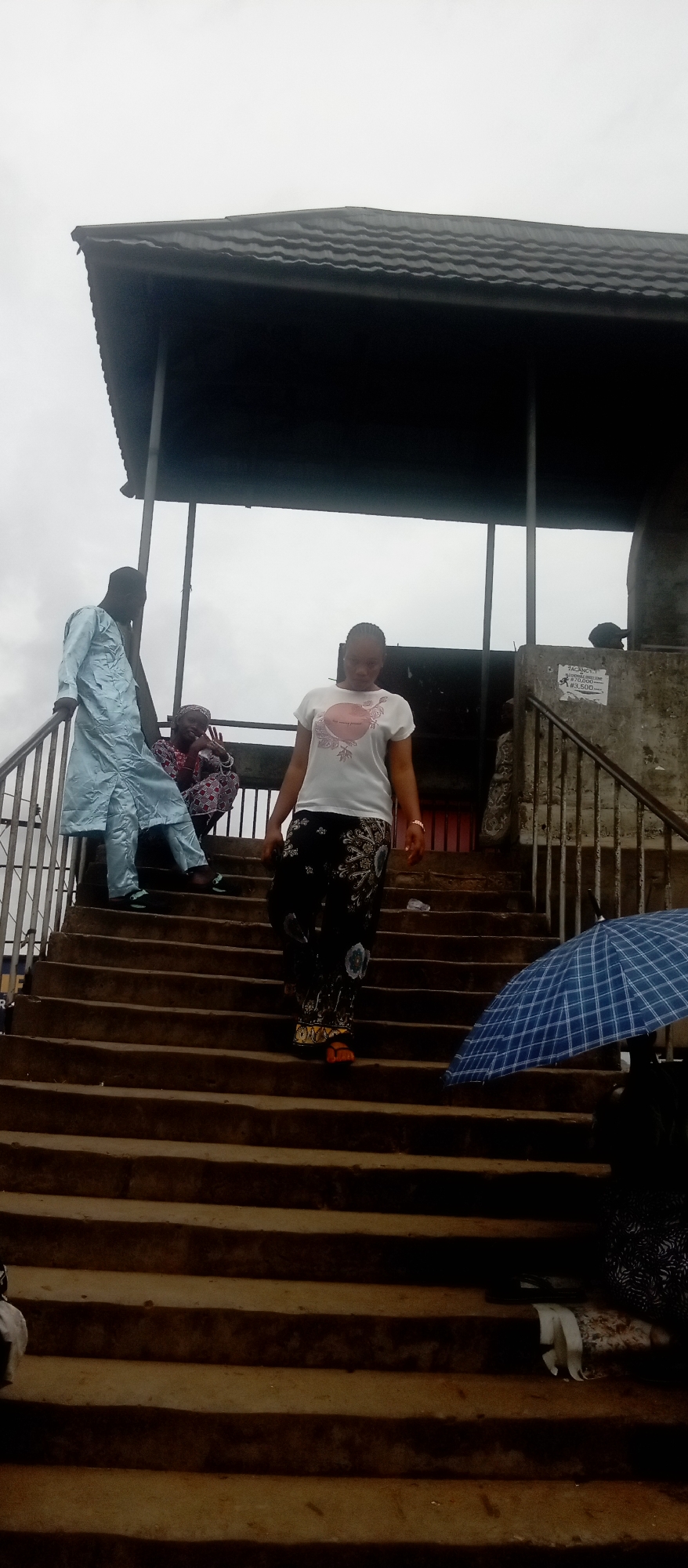 The image shows three people on a concrete stairway in Idemili, Nigeria, with a shelter overhead. A woman in a white t-shirt and patterned pants is descending the stairs. To her left, a man in a light blue robe stands on a higher step, and a woman wearing a patterned headscarf sits near him. A blue and white checkered umbrella is visible on the lower right. A poster is visible on the side of the stairs. The sky is overcast.