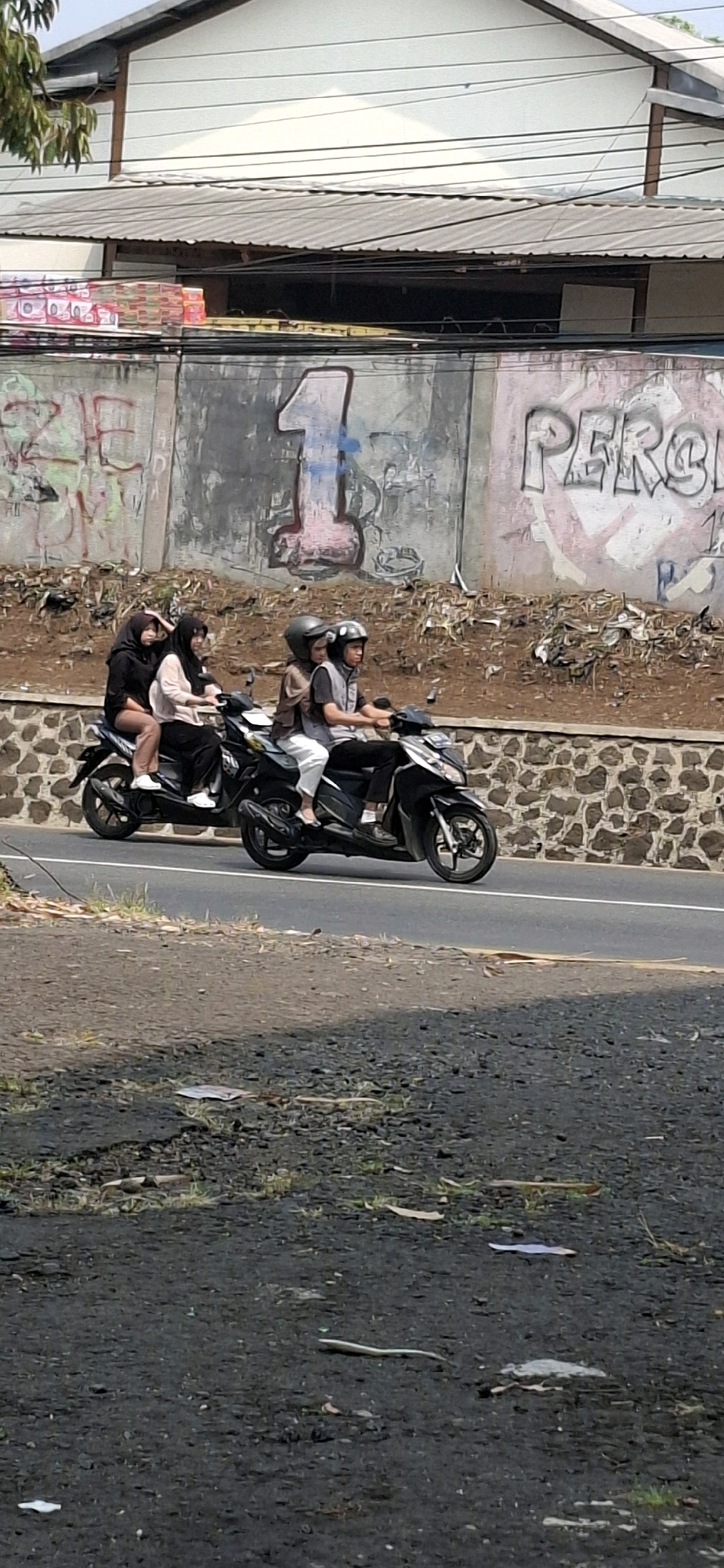 In the city of Cilaku, Indonesia, a scene unfolds on a paved road bordered by a stone retaining wall and a sloped embankment with scattered debris. Two motorcycles, each carrying two people, are captured in motion. The first motorcycle, on the left, transports three individuals. Two women are seated behind the driver, both wearing hijabs; one in a black hijab and the other in a pinkish-beige hijab. The woman in the black hijab is positioned at the rear, while the woman in the pinkish-beige hijab is in the middle. The third person, whose attire is not fully visible, is seated in front of them, presumably driving. The second motorcycle, to the right, carries two individuals. The driver is wearing a helmet and a gray vest over a dark shirt, with white pants. A passenger sits behind the driver, also wearing a helmet. Both motorcycles are moving from left to right across the frame. Behind the motorcycles, a concrete wall serves as a backdrop, adorned with graffiti. Prominent among the graffiti are a large number "1" in pink and a stylized word that appears to read "PERCA" in pink and white. Above the wall, a building with a white corrugated metal roof and siding is visible, along with a tangle of overhead wires. The foreground of the image consists of a rough, dark-colored surface with scattered stones, debris, and some sparse vegetation. The lighting suggests it is daytime, casting shadows across the scene.