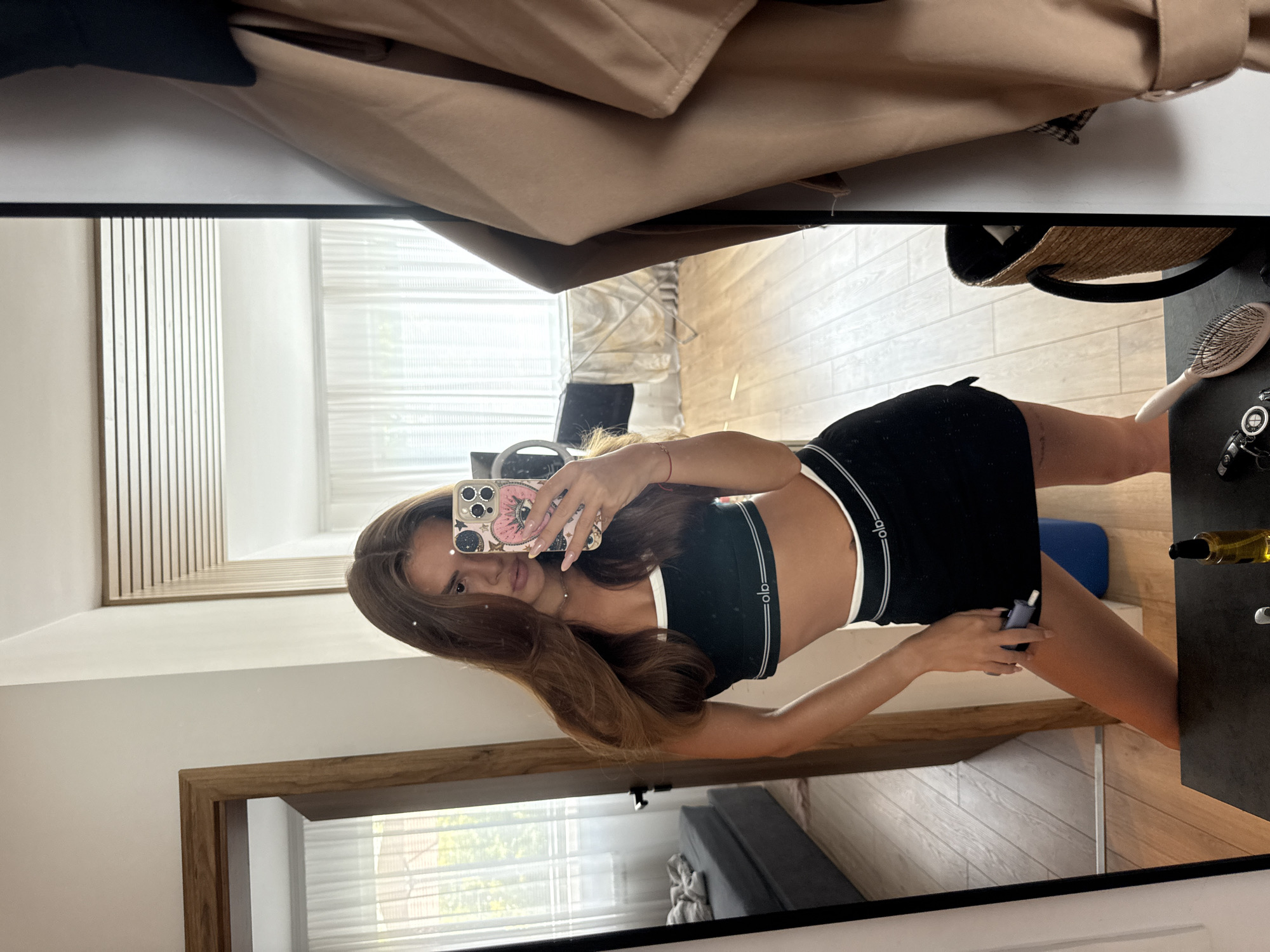 A young woman is taking a mirror selfie in a well-lit room. She is wearing a black sports bra and a black mini-skirt with white trim. Her long, brown hair is down, and she is holding a smartphone in her right hand, capturing her reflection. The background reveals a doorway to another room on the left, showing a sofa and a window with sheer curtains. On the right, a beige trench coat is hanging, and a woven basket is visible. The room has wooden floors and a white ceiling with horizontal wooden accents. A table in the foreground holds a bottle of oil and a hairbrush. The overall setting appears to be an apartment. This scene is not specifically located in the city of Plovdiv, Bulgaria, as no identifying landmarks or contextual information suggest this location.