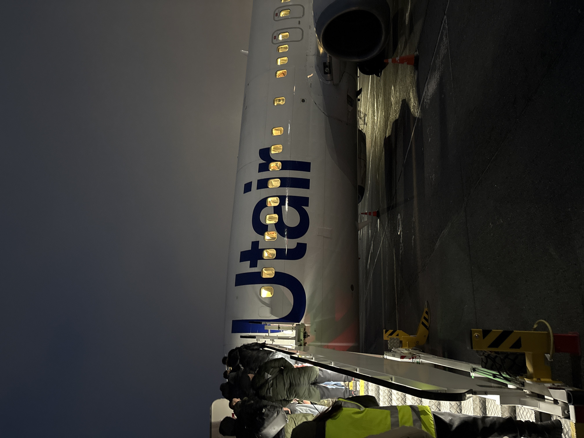 The image depicts an UTair passenger plane at night, likely at an airport in Nizhnevartovsk, Russia. Passengers are disembarking via a mobile stairway, with ground crew visible in reflective vests. The plane's fuselage, marked with "UTair" in large blue letters, is illuminated by interior lights. The engine is visible along with orange cones next to the plane. The surroundings appear to be dark and overcast.