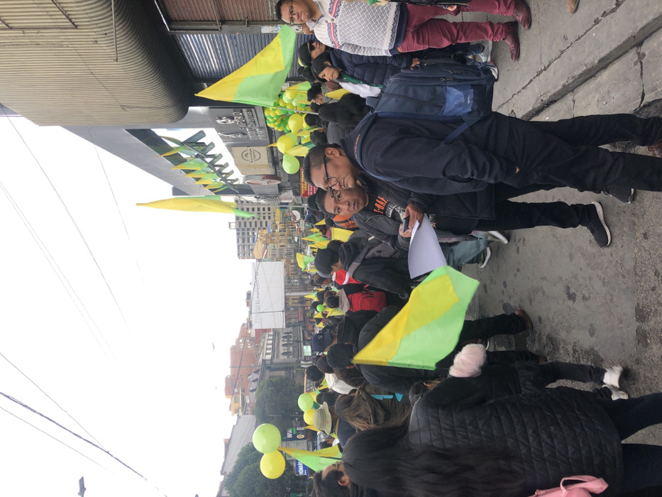 A street protest unfolds with participants holding yellow and green flags and balloons. The crowd, dressed in various clothing, fills the street, walking between buildings and under awnings. Some individuals carry papers. The sky appears overcast, suggesting a cloudy day.