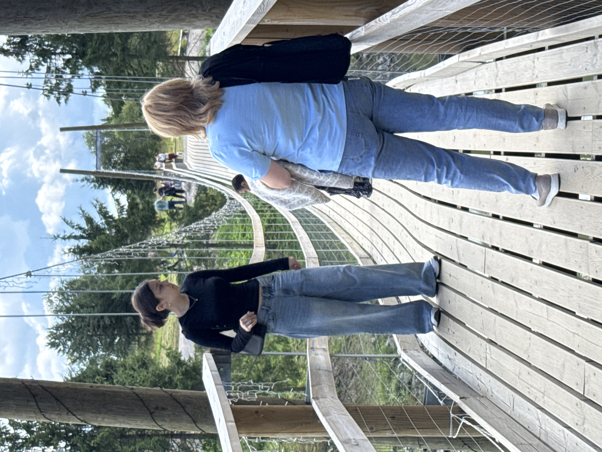 Two women are walking on a suspension bridge made of wooden planks. The woman in the foreground is wearing a light blue t-shirt and blue jeans. She has blonde hair and a black backpack slung over her right shoulder. She is holding a patterned blanket in her left arm. The woman in the background is wearing a black long-sleeved shirt and wide-leg blue jeans. She has dark brown hair pulled back and is carrying a small dark purse on her left shoulder. The bridge is surrounded by lush green trees and a cloudy sky. Other people can be seen walking on the bridge in the distance. The location is described as a suspension bridge in the city of Khankanmyaki, Russia.