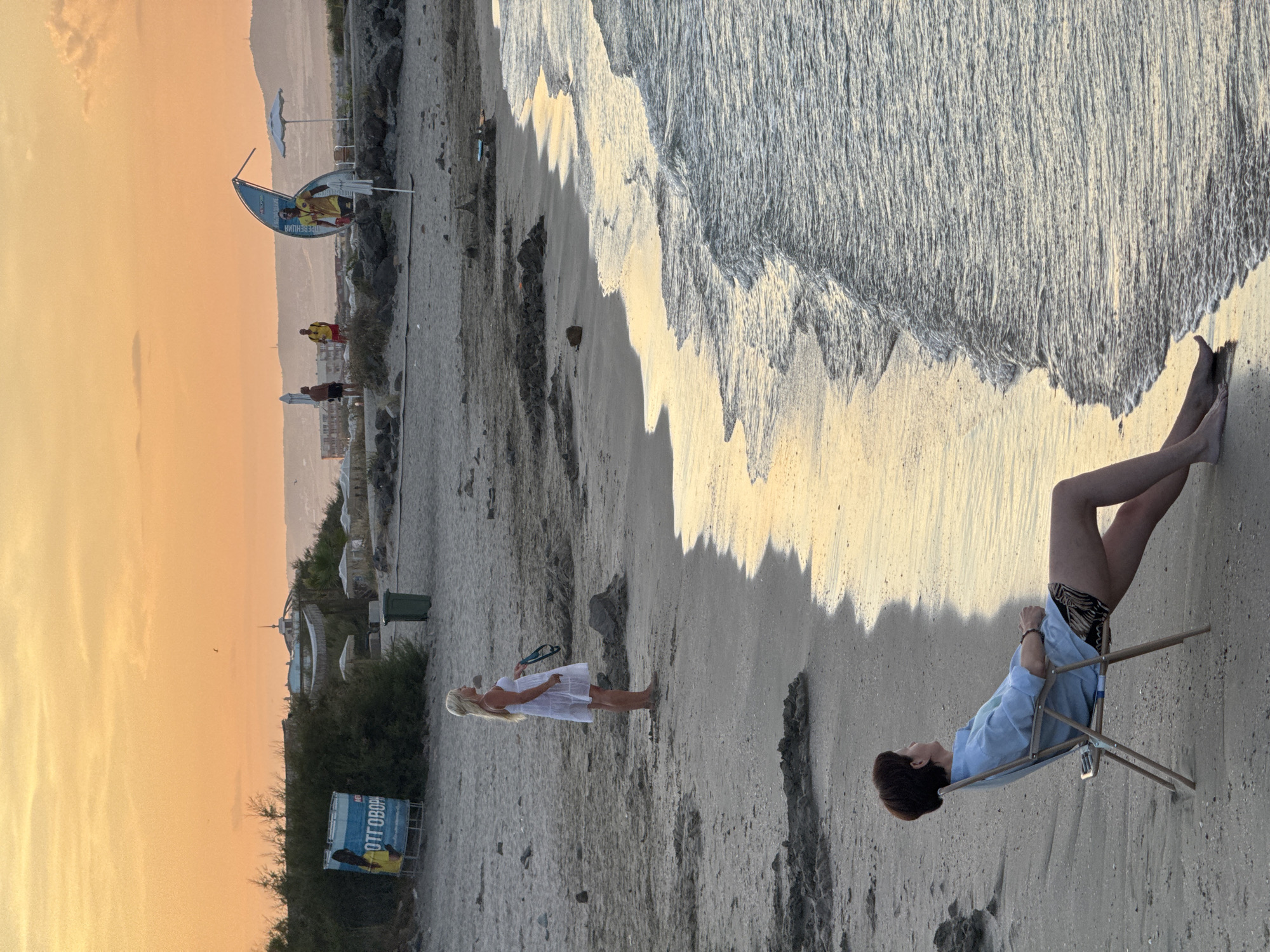At dusk on a beach in Pomorie, Bulgaria, a woman in a white dress stands facing the sea. Further up the shore, another woman sits in a folding chair, looking out at the water. In the background, several beach umbrellas and structures are visible, along with a few people. The sky is a warm orange and yellow, indicating the sunset. The ocean waves are gently lapping at the sandy shore.