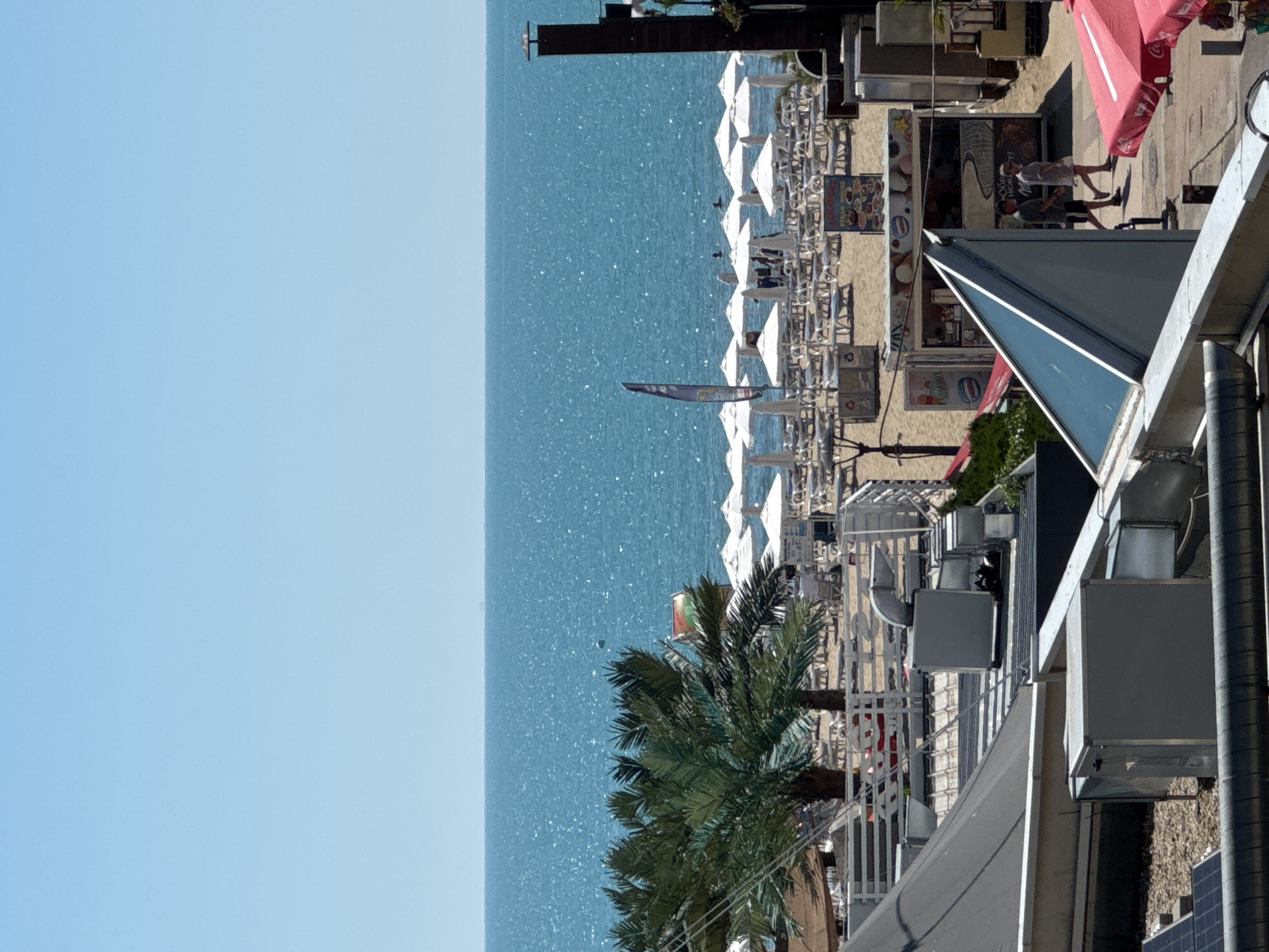 The image shows a sunny day at a beach resort. In the foreground, there are rooftops with ventilation systems and solar panels. Moving towards the background, a palm tree is visible in front of rows of white beach umbrellas and lounge chairs. The ocean is in the distance, with a clear blue sky above. Several small kiosks or food stalls are set up on the beach. Two people are walking on a path near the stalls. The overall impression is of a lively, popular beach destination.