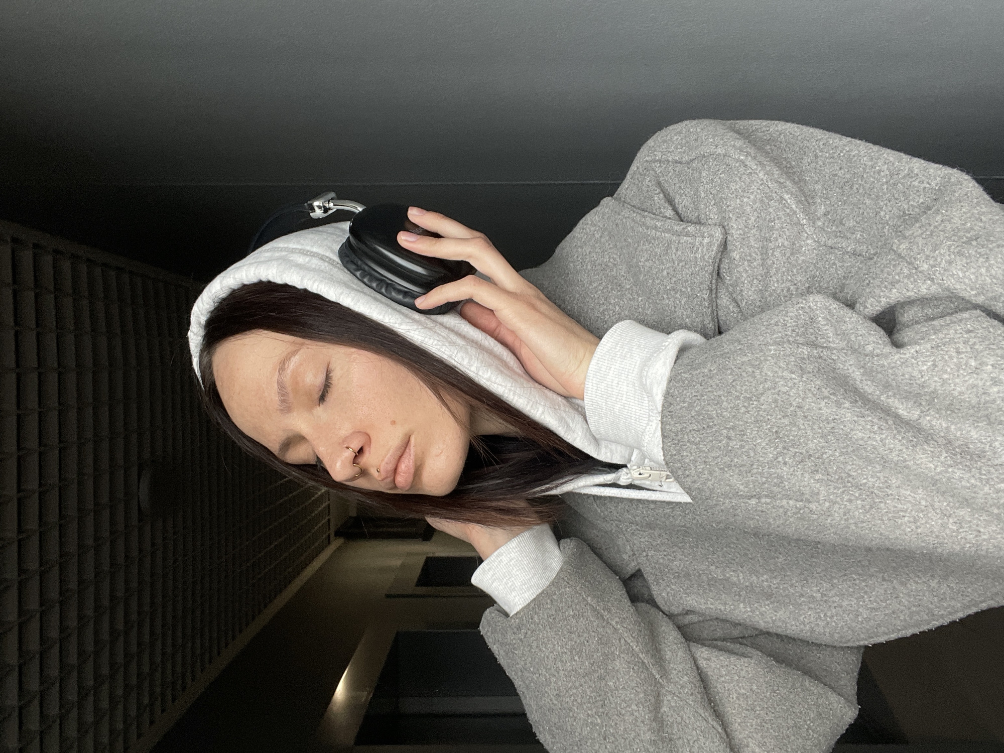 A woman is pictured wearing a white hoodie, grey coat, and black headphones. Her eyes are closed. The background appears to be a building interior with a dark wall. The location of the image is unknown, and there is no information to suggest it was taken in Izhevsk, Russia.