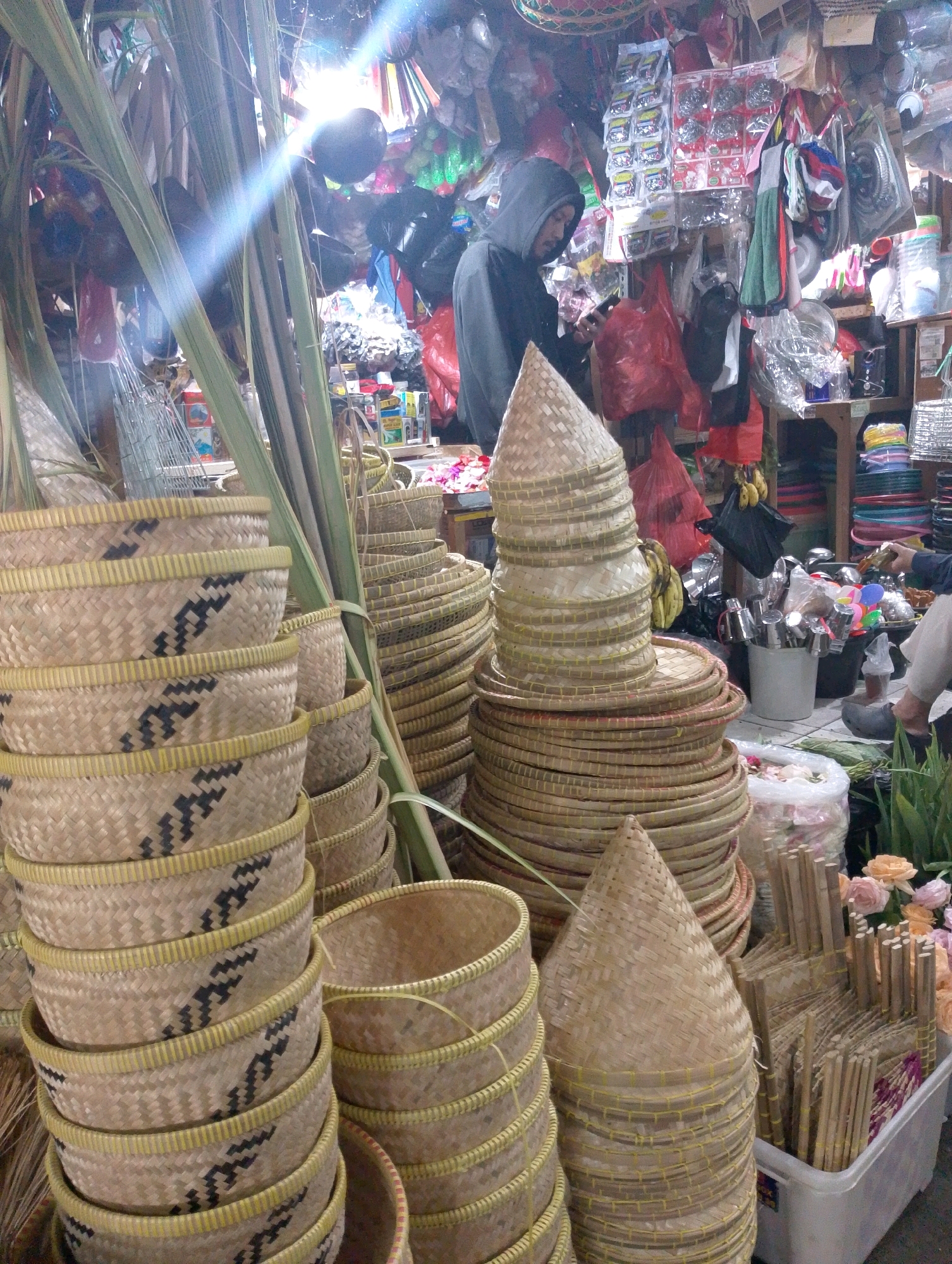The image depicts a bustling street-side stall in Lembang, Indonesia, likely a local market. The foreground is dominated by an abundant display of woven bamboo products. Stacked high are numerous round, shallow baskets with intricate black and yellow woven patterns. Several conical-shaped woven hats, also made of bamboo, are stacked in a prominent position. Behind these, more woven items are visible, including a large, tiered stack of flat, round woven trays. In the background, the stall is crowded with a variety of goods. A man wearing a black hoodie and a face mask is visible, appearing to be looking at his phone. Other items for sale include plastic bags, packaged goods, kitchen utensils in a bucket, and colorful stacks of plastic plates. The stall is also adorned with greenery, including long, thin palm-like leaves. Sunlight streams down, creating bright glares and casting shadows, suggesting it is daytime. The overall impression is one of a vibrant, well-stocked market stall with a diverse range of handmade and manufactured items.