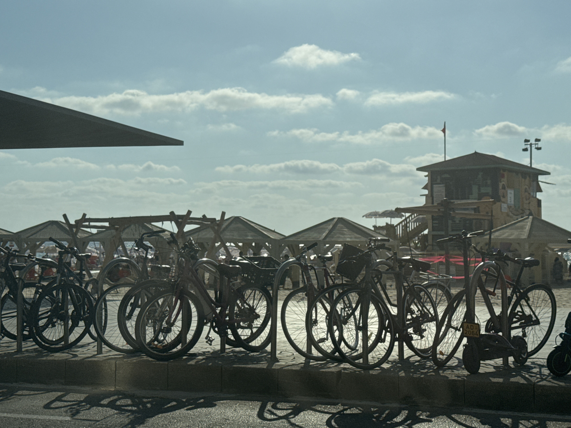 The image depicts a row of bicycles parked along a sidewalk with a paved surface. In the background, there are beach structures with thatched roofs and a lifeguard tower, suggesting a seaside location. The sky is partly cloudy. The bicycles are silhouetted against the bright sky, making it difficult to discern specific details about their models or colors. The overall impression is that of a public amenity area, likely a beach or promenade in Tel Aviv, Israel, providing access to the beach via bicycle.
