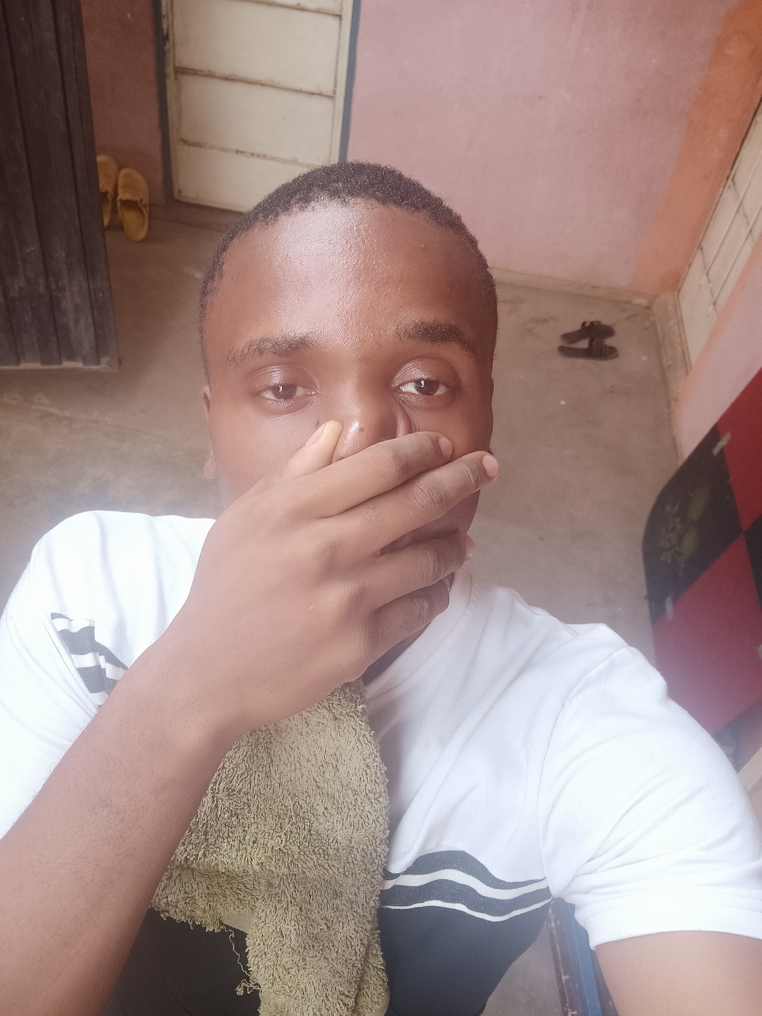 The image shows a man in an indoor setting in Narayi, Nigeria. He is covering his mouth with his right hand and holding a light green towel with his left hand. He is wearing a white T-shirt with black stripes on the sleeves. Behind him, there's a closed door with a window, a pair of yellow sandals, and a pair of black sandals. The walls appear to be painted in a combination of pink, orange, and red. The floor is concrete.