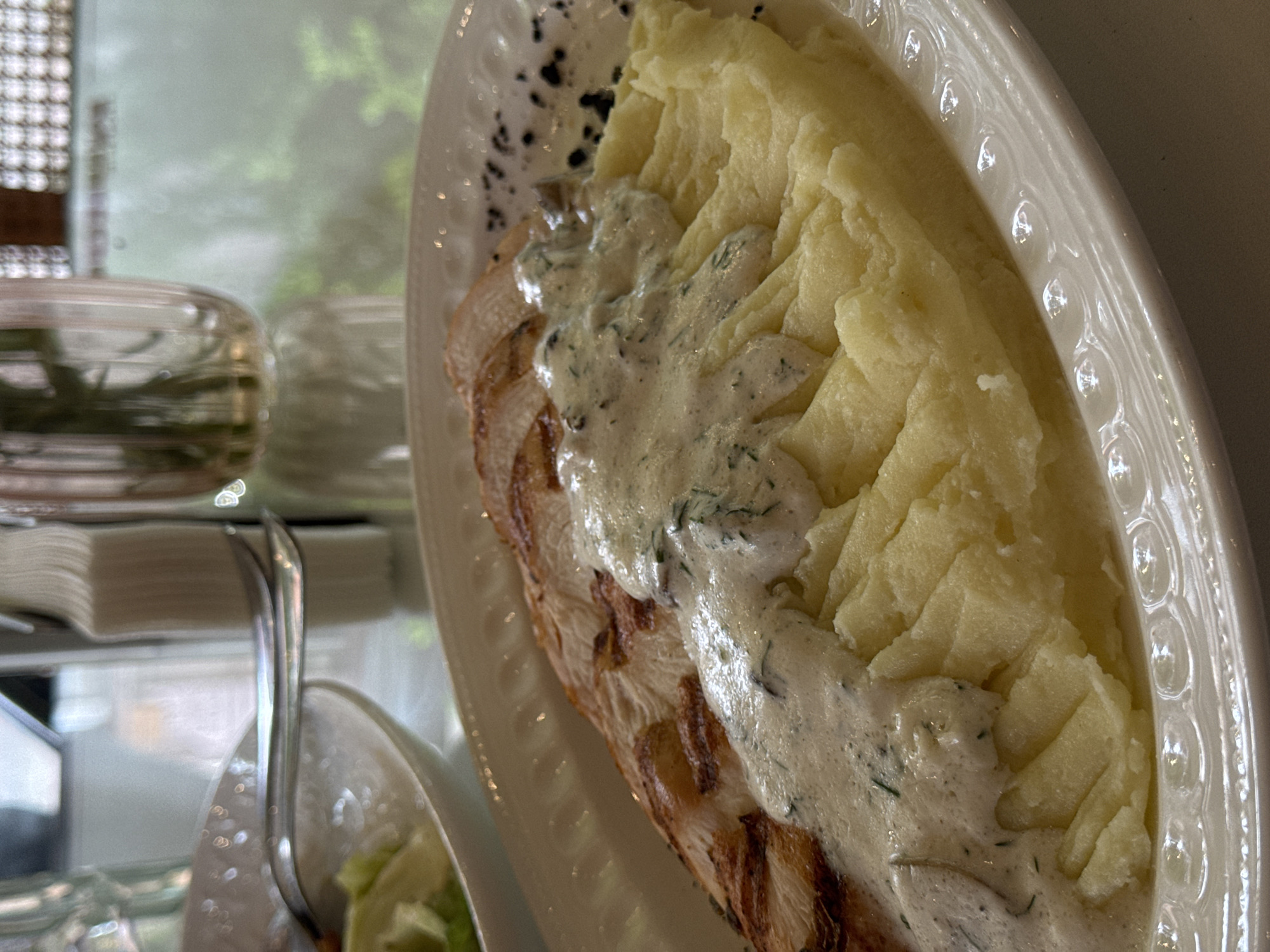 A white oval plate holds a meal consisting of sliced, grilled chicken topped with a creamy, dill-flecked sauce, alongside a portion of mashed potatoes. The plate has a decorative edge. The scene is in Yerevan, Armenia. In the background, there's a blurry view through a window, showing greenery. To the left of the plate, out of focus, are a metal serving utensil and a partially visible side dish of salad in a clear bowl. A glass container with sprigs of herbs sits on a surface next to a stack of white napkins.