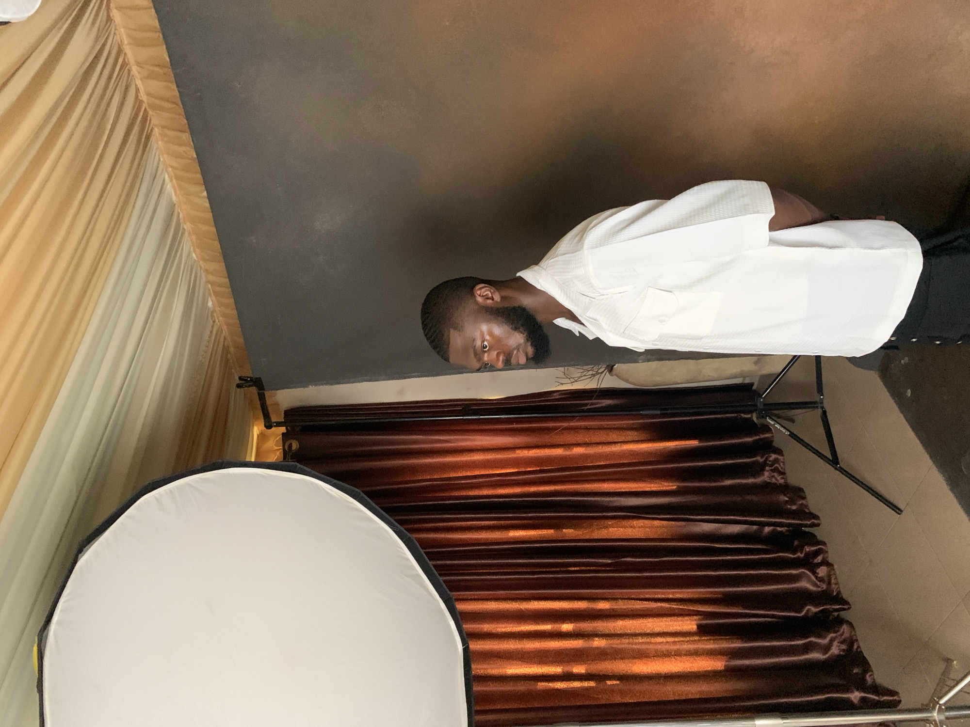 The image shows a man with dark skin and a beard, wearing a white collared shirt. He is positioned in a studio setting, with a dark brown backdrop behind him. To the left of the man, there are beige curtains with vertical pleats. In the foreground, a large white softbox, a piece of photography equipment, is visible on the left side of the frame. To the right of the softbox and behind the man, there is a brown, textured material with horizontal folds, possibly a backdrop or draped fabric. A tripod stand for studio lighting is also visible to the right, behind the man. The man's head is turned to his left, and he appears to be looking down. The lighting suggests an indoor studio environment. There is no visible text in the image. The scene does not provide any cues about the time of day or weather.