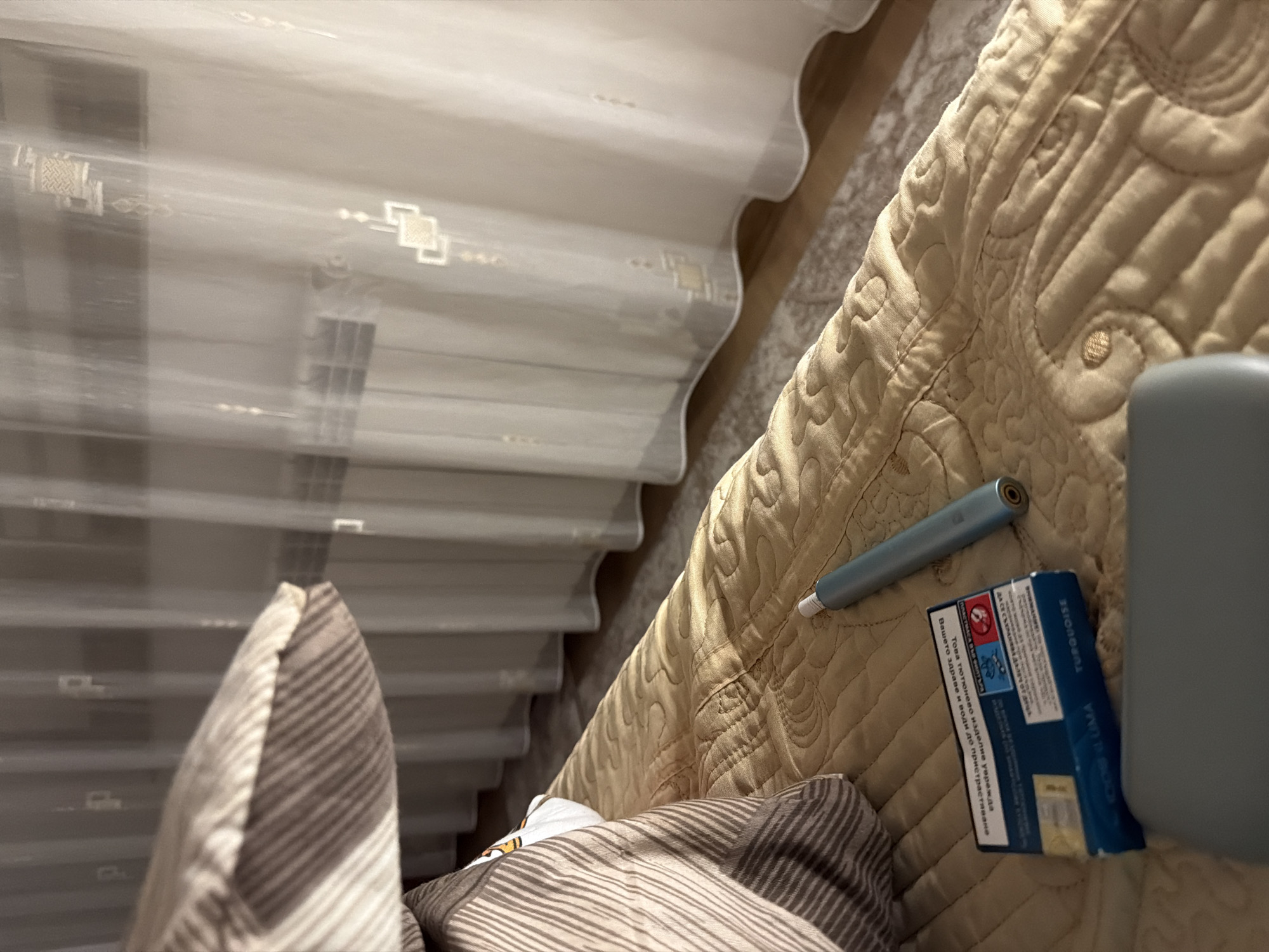 An indoor scene, likely a bedroom, is captured from a low angle. In the foreground, a light brown, quilted blanket covers a bed. On the blanket, to the left of center, is a partially visible pack of turquoise cigarette-like cartridges, with Russian text on the packaging. Next to the cartridge pack, to the right, lies a slender, light blue electronic smoking device with a metallic end. A striped pillow is visible in the upper left corner of the image. In the background, sheer white curtains hang, obscuring a window and a radiator. The overall lighting suggests an evening or dimly lit indoor environment. The image does not provide information about the city of Zmeitsa, Bulgaria.