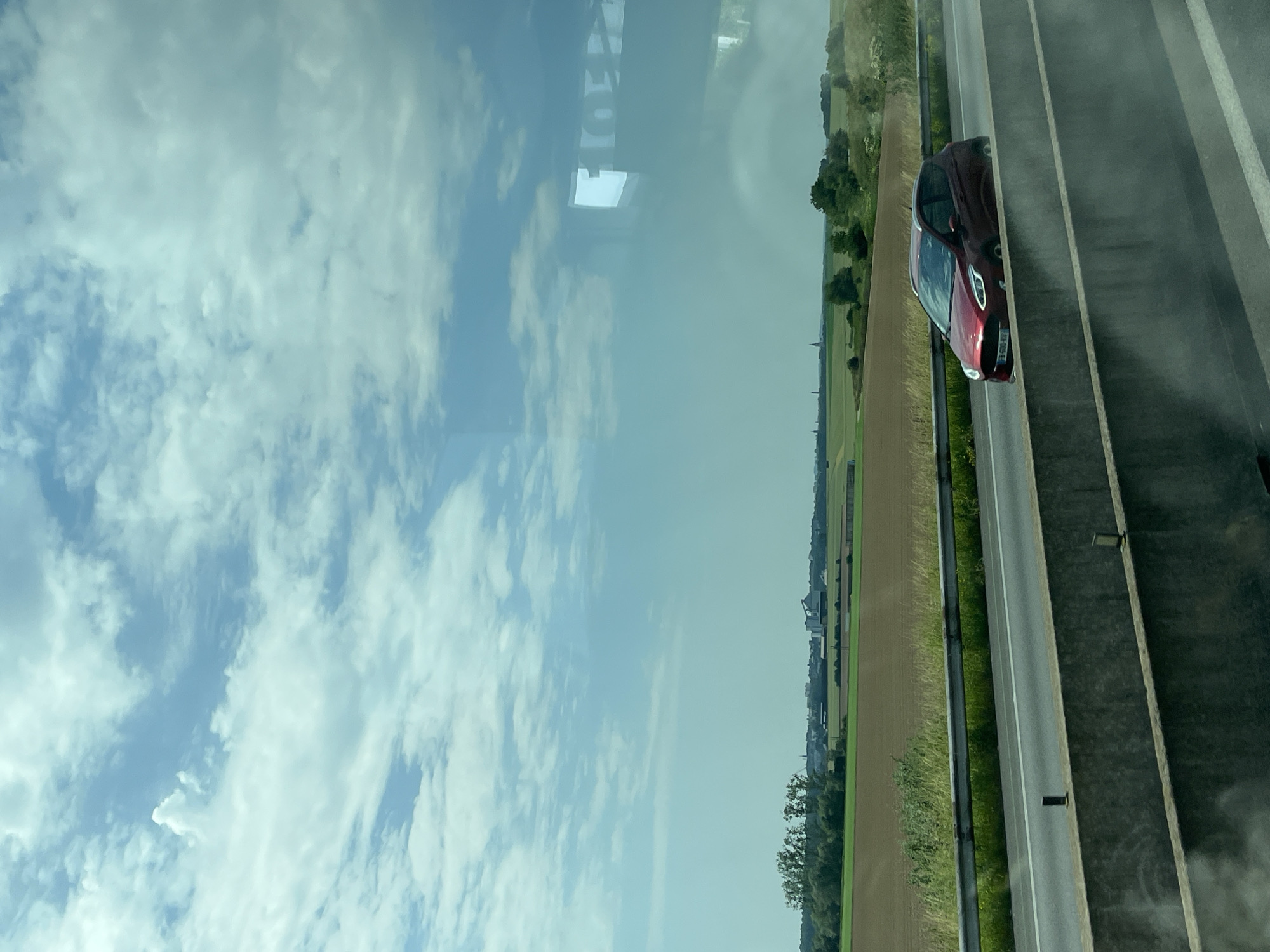 The image depicts a highway scene in Eswars, France. In the foreground, a concrete barrier runs horizontally across the bottom of the frame. Beyond the barrier, a red hatchback car is driving on the highway. The car has its headlights on and appears to be moving at a moderate speed. The highway is flanked by a grassy verge and a metal guardrail.
In the middle ground, a vast agricultural field, predominantly brown and green, stretches towards the horizon. A line of trees separates the field from the highway. In the distance, a cluster of buildings, including what appears to be a silo or industrial structure, are visible, along with several church spires suggesting a small town or village.
The background is dominated by a bright blue sky with scattered white clouds, characteristic of a sunny day. There are reflections on the surface from which the photo was taken, showing geometric shapes and light. The overall impression is a snapshot of rural French landscape viewed from a moving vehicle.