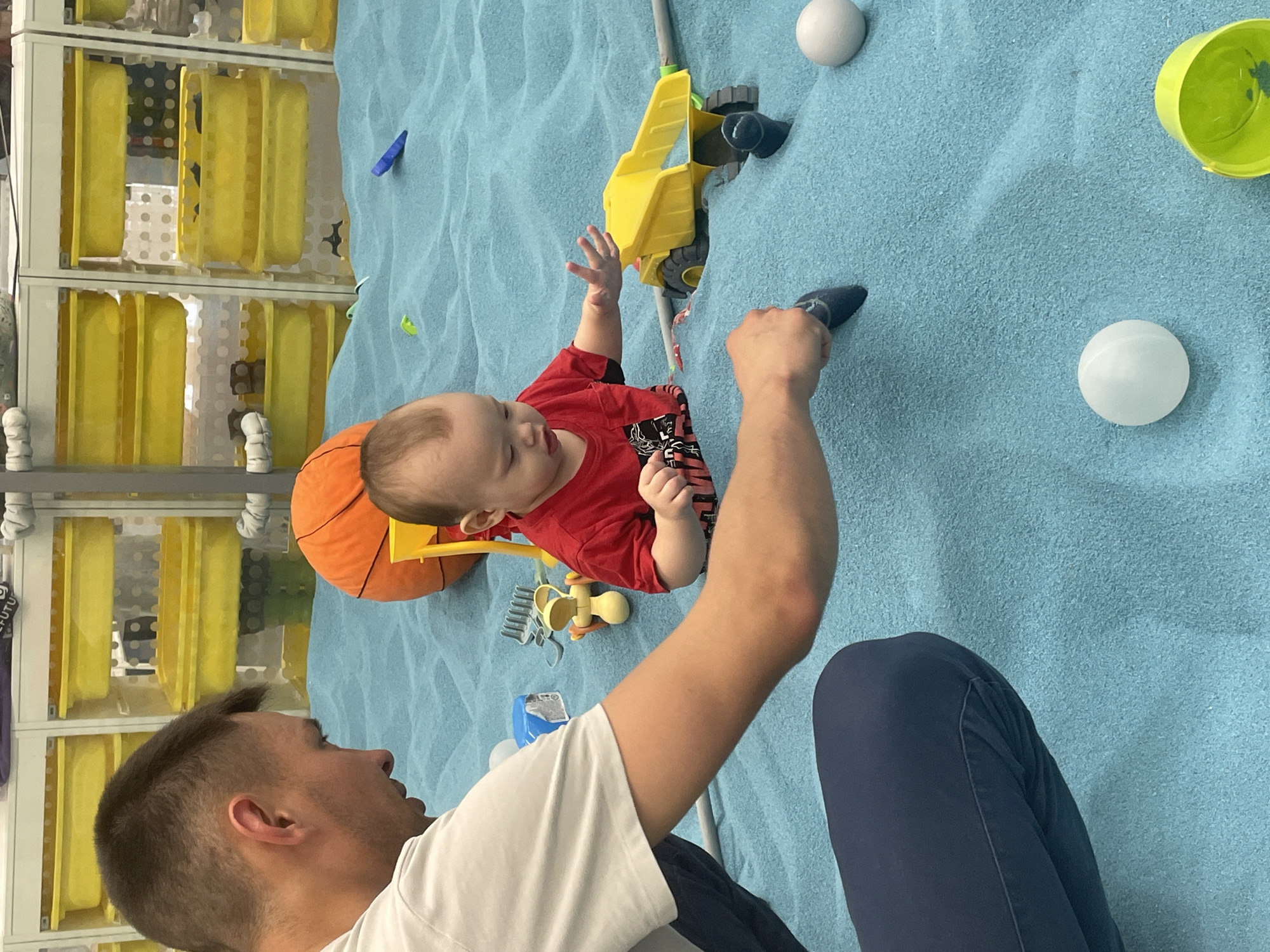 A man and a baby are playing on a light blue textured surface. The man is on the left, wearing a white t-shirt and blue pants. He has short brown hair. The baby is in the center, wearing a red shirt. Several toys are scattered around: a yellow toy truck attached to a gray bar, an orange basketball, a yellow windmill toy, a green bucket, and two white balls. In the background, on the left, are shelves with yellow containers. The location is implied to be in the city of Perm, Russia, but this cannot be verified from the image.