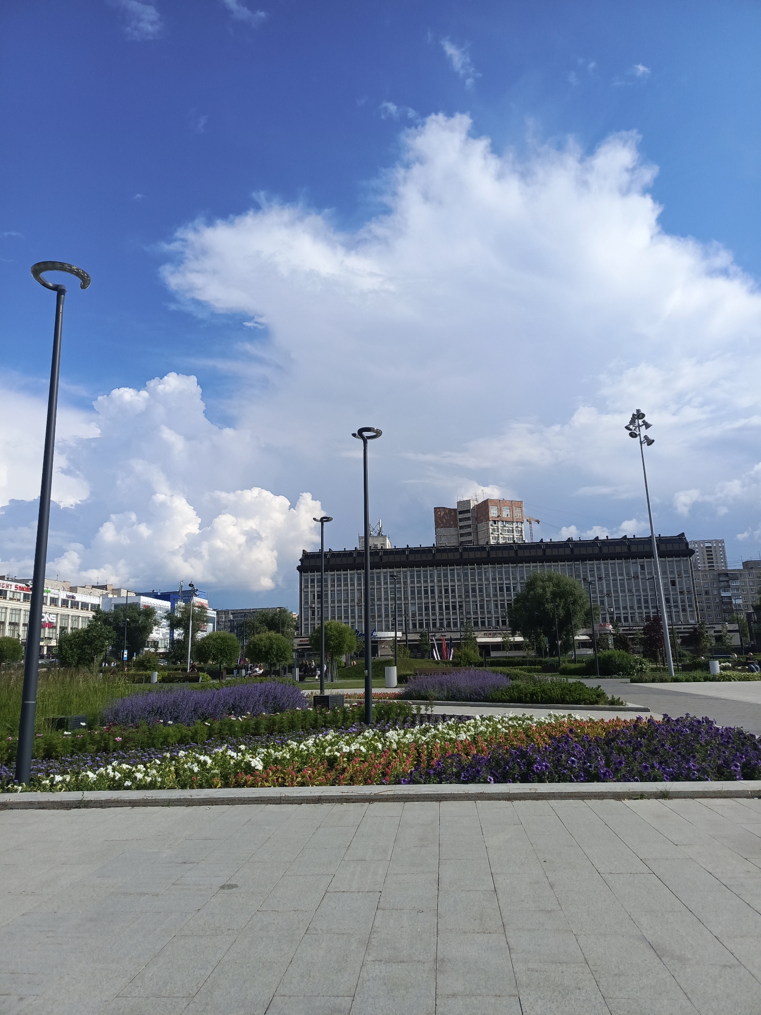 The image depicts a wide, paved public square in front of a large, modernist building. The square features a colorful flowerbed with rows of purple, white, and orange flowers, bordered by greenery and grass. Two tall, dark lampposts stand on either side of the foreground, and a third, more elaborate lamppost with multiple lights is visible further back.
In the midground, the large building dominates the scene. It appears to be an office or administrative building, characterized by a grid of windows and a darker, horizontal band along its roofline. Some construction or renovation work is visible on top of a section of the building, with scaffolding and a crane. Adjacent to this main building are other, slightly less imposing structures, including some with visible signage and trees.
The sky is a vibrant blue, with large, fluffy white cumulus clouds that suggest fair weather, possibly with a chance of rain later. The overall impression is one of a well-maintained urban public space on a clear day.
Based on visual cues and architectural styles, the scene is likely located in the city of Perm, Russia, specifically on the Esplanade (also known as the "Olimpiyskaya Naberezhnaya" or Olympic Embankment), a central public space in the city. The large building in the background is recognizable as the Perm Academic Theatre-Theatre building, a prominent landmark.