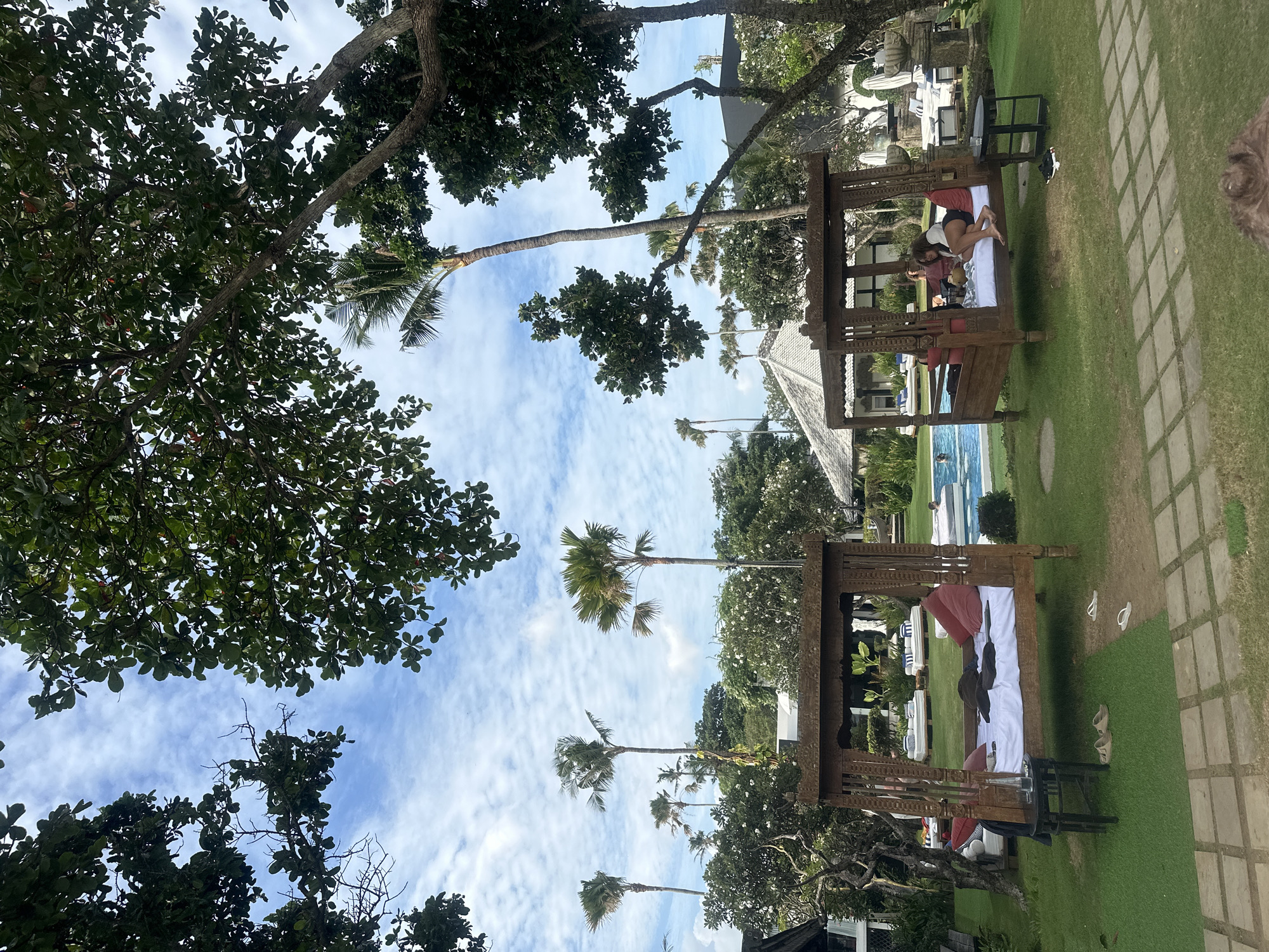 The image depicts an outdoor scene in North Kuta, Indonesia, featuring two wooden, canopy-style daybeds on a green lawn. One daybed is occupied by two individuals, one appearing to be reclining while the other is in a seated position. Behind the daybeds, a blue swimming pool is visible. Palm trees and various other trees dot the landscape. A paved walkway with square stones runs adjacent to the lawn. The sky above is blue with scattered clouds. The scene suggests a resort or vacation setting.