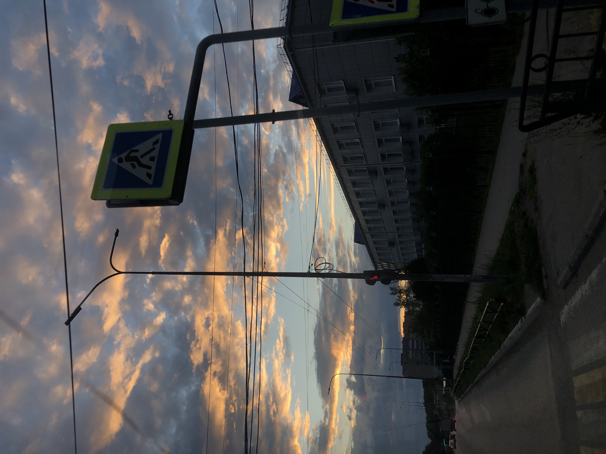 The image depicts a street scene in Chaikovsky, Russia, during a dramatic sunset. The sky is filled with a mixture of dark, stormy clouds and bright, illuminated clouds tinged with orange and pink hues. In the foreground, a crosswalk is visible on the asphalt road, marked with yellow and white stripes. To the right of the crosswalk, a tall street light pole stands, topped with a traffic signal displaying a red light. A pedestrian crossing sign, featuring a blue square with a white triangle and a silhouette of a person walking, is mounted on a pole to the right of the street light.
Further down the street, a modern, multi-story building with a gray facade and numerous windows is visible. The building is flanked by trees and a dark metal fence. Overhead, a network of electrical wires crisscrosses the sky, silhouetted against the colorful clouds. In the distance, more buildings and trees are faintly discernible, suggesting a suburban or urban environment. A few cars are seen on the road, their headlights on, indicating a low-light condition. The overall impression is one of a typical evening in a Russian city, marked by the striking beauty of the sunset.