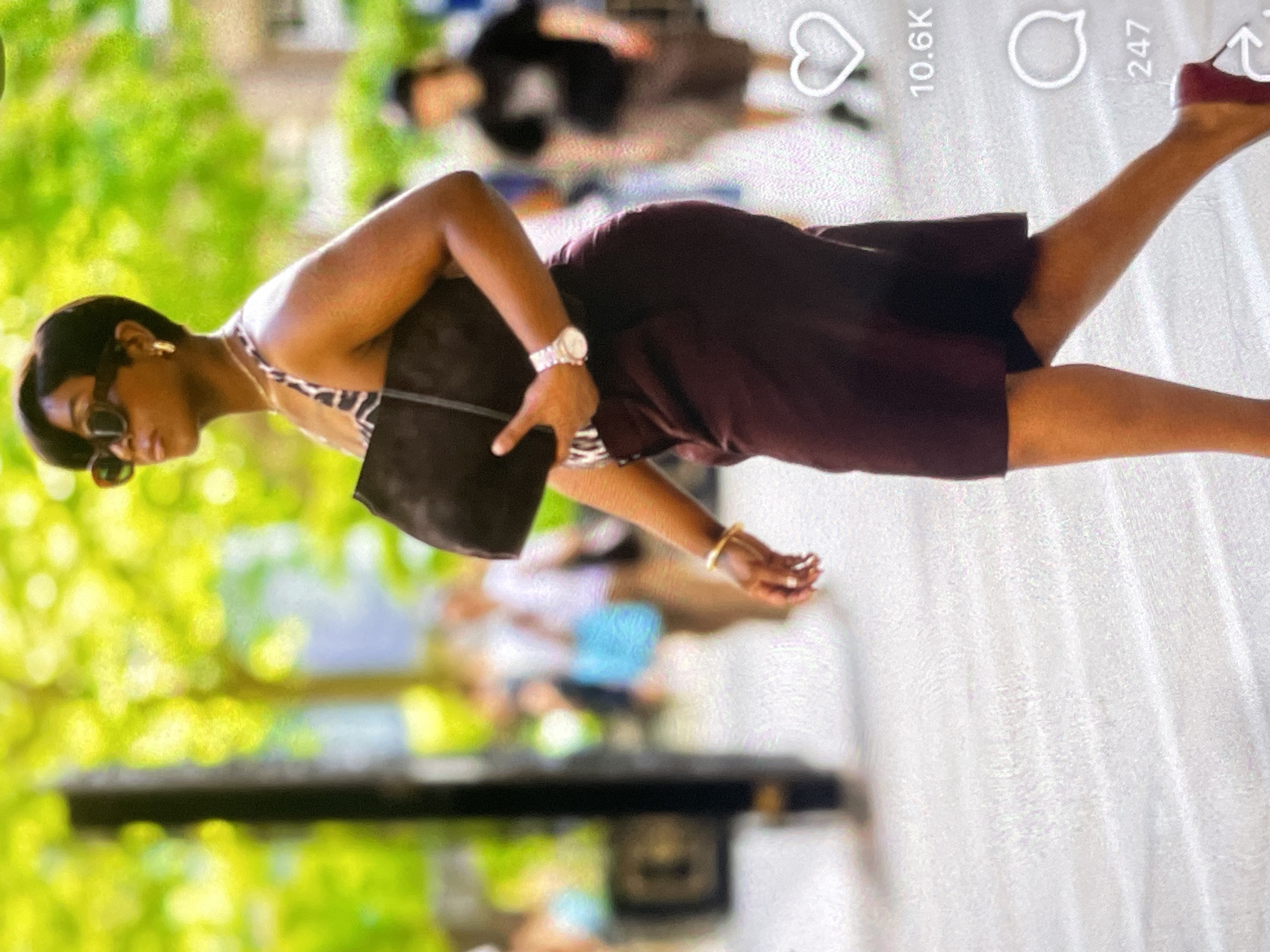 A medium shot captures a Black woman from the knees up, walking outdoors in a city setting. She has a short, dark hairstyle, wears large, dark sunglasses, and large hoop earrings. Her outfit consists of a patterned halter top and a dark red skirt. She carries a dark, oversized clutch bag in her left arm. On her left wrist, she wears a silver watch. A gold bracelet adorns her left wrist, and a ring is visible on her left hand. Her right hand is extended forward, slightly bent at the elbow. Her legs are crossed as she walks, with her right leg slightly in front of her left. She wears dark red high-heeled shoes. The background is blurred, showing a bright, sunlit environment with greenery, likely trees, and other pedestrians in motion, suggesting a bustling urban scene. A tall, dark vertical object, possibly a pole or a column, is visible on the left side of the frame. Social media engagement metrics, including a heart icon with "10.6K" and a speech bubble icon with "247," are overlaid on the right side of the image.