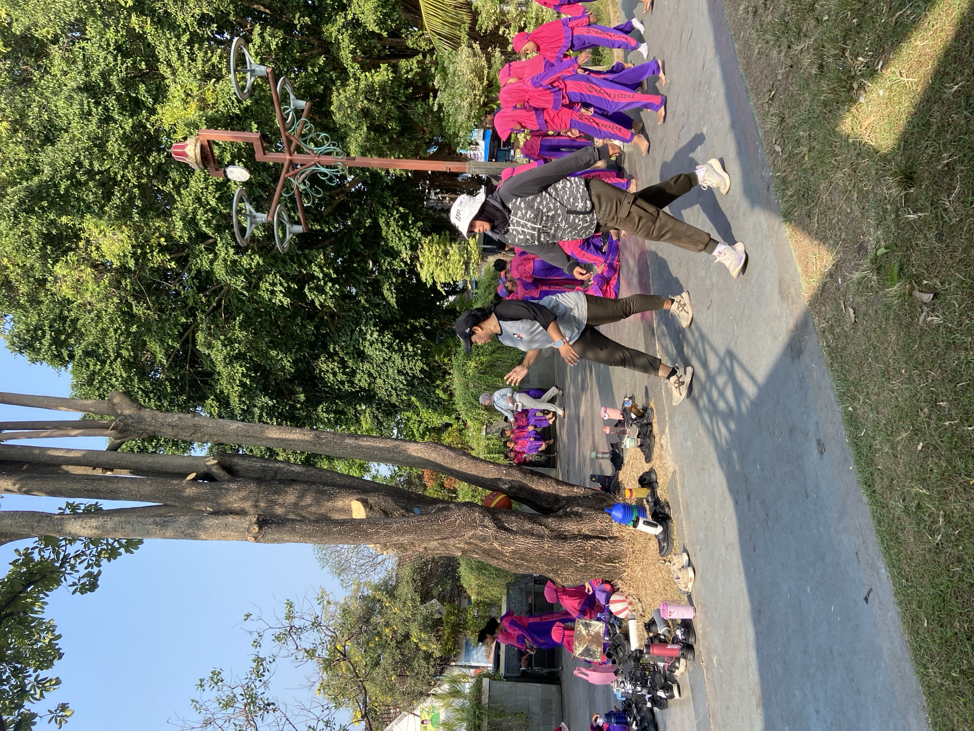 The image shows an outdoor scene likely in Kecamatan Pati, Indonesia, with a group of children and adults near a large tree. The children, predominantly wearing bright pink and purple outfits, appear to be participating in an organized activity, possibly a school event or performance. Some are seated on benches, while others are standing or walking. There is a significant collection of footwear and water bottles gathered at the base of the tree, suggesting a break or preparation for an activity. Two adults are in the foreground, one wearing a grey hooded jacket and a white bucket hat, and another wearing a black cap and grey long-sleeved shirt. The environment includes lush green trees and a blue sky, indicating it's daytime. A lamppost with decorative elements is visible in the background.